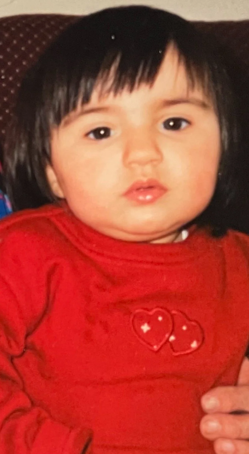 A young child with black hair, wearing a red shirt with two hearts and stars embroidered on it, sitting on a brown chair.
