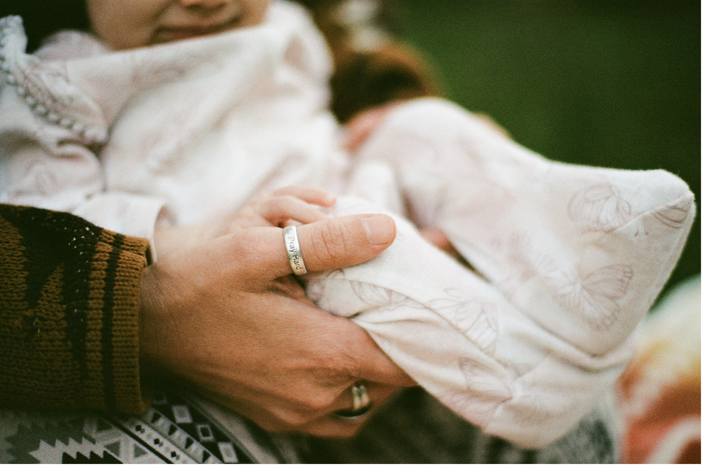 Person holding a baby outdoors, with focus on the baby's hand and the person's hand, which has a ring and a ringband that says 'Pray Hard'.