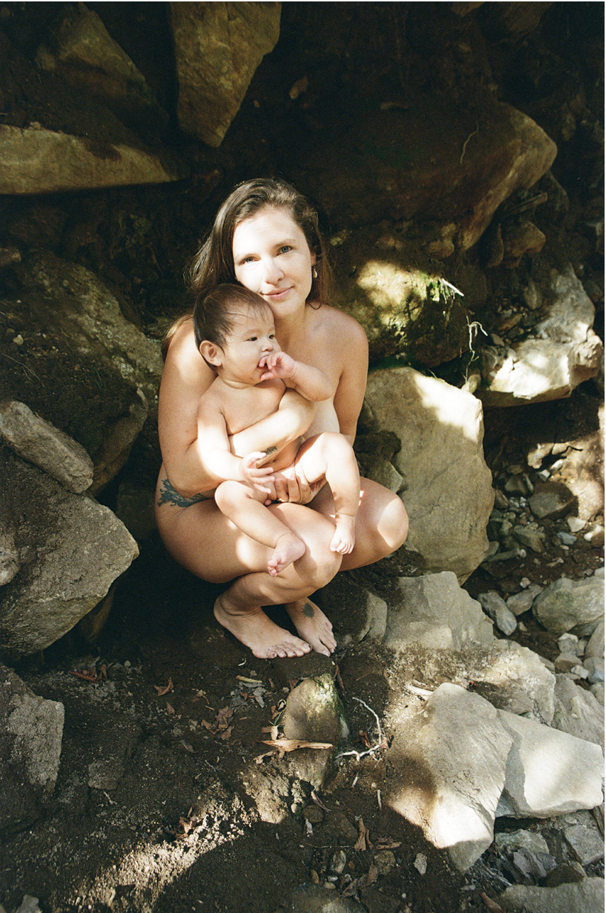 A woman with long brown hair sitting on rocks outdoors, holding a young child with short dark hair. The woman has a tattoo on her left side and is looking at the camera, while the child is sucking their thumb and looking to the left. They are in a natural setting with large rocks and dirt around them, and sunlight filtering through trees.