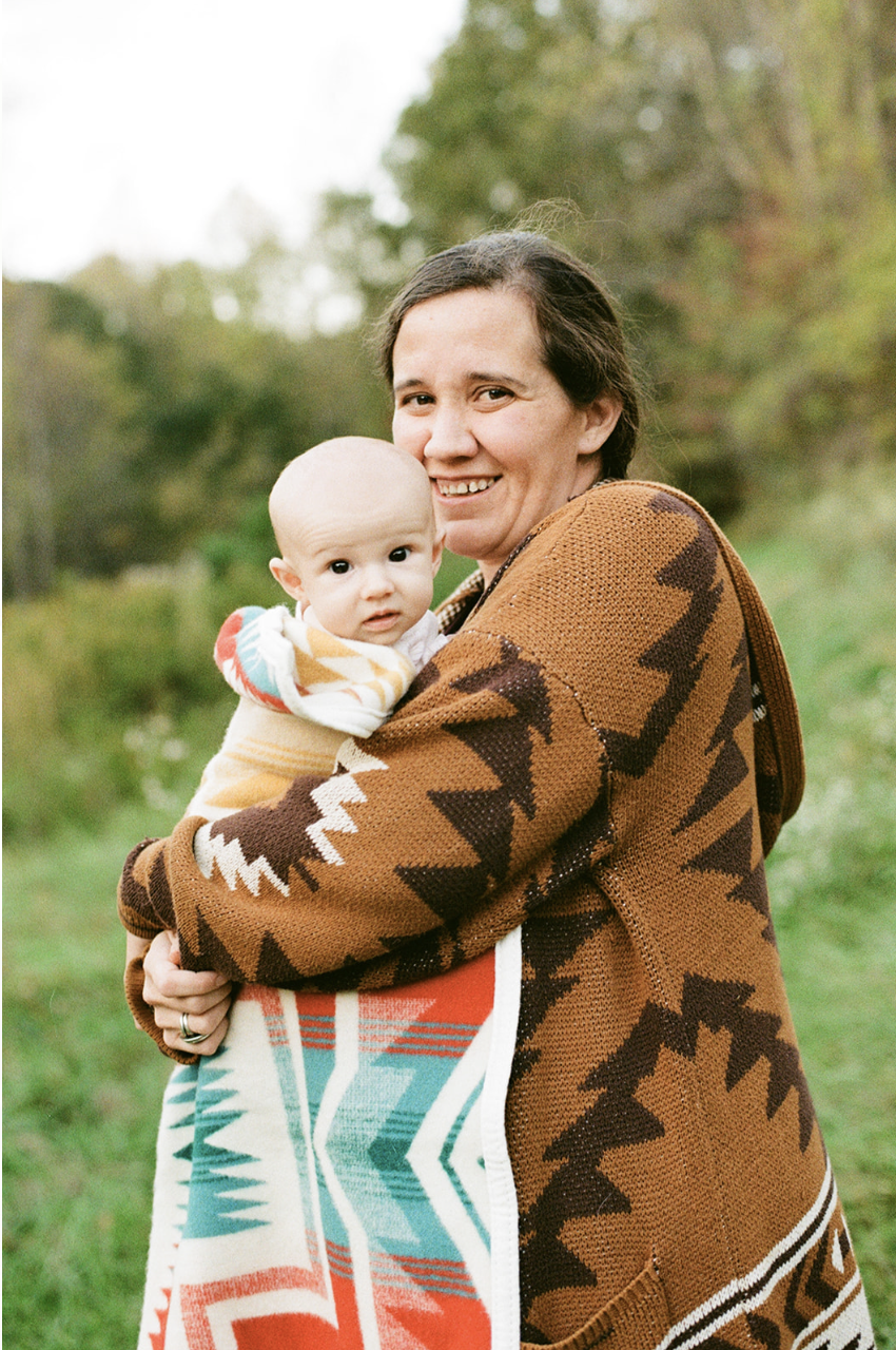 A woman holding a baby outdoors in a grassy area with trees in the background, both dressed in patterned sweaters.