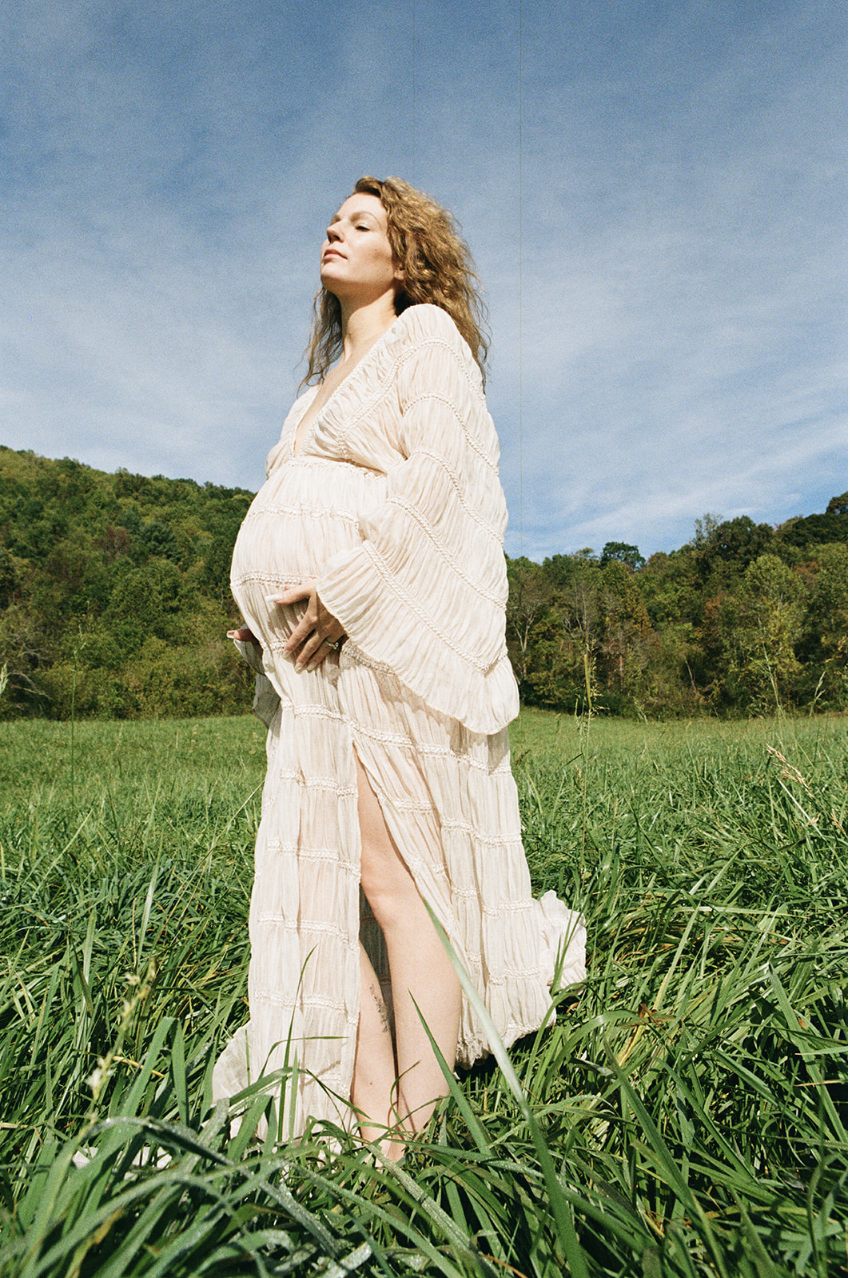 A pregnant woman standing in a grassy field, wearing a flowing cream-colored dress with a slit, with a backdrop of green trees and a blue sky.