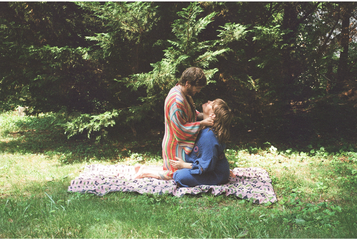 Two women on a pink blanket in a forest, one with short hair in a multicolored striped dress, and the other with long hair in a blue dress, engaging in an intimate moment.