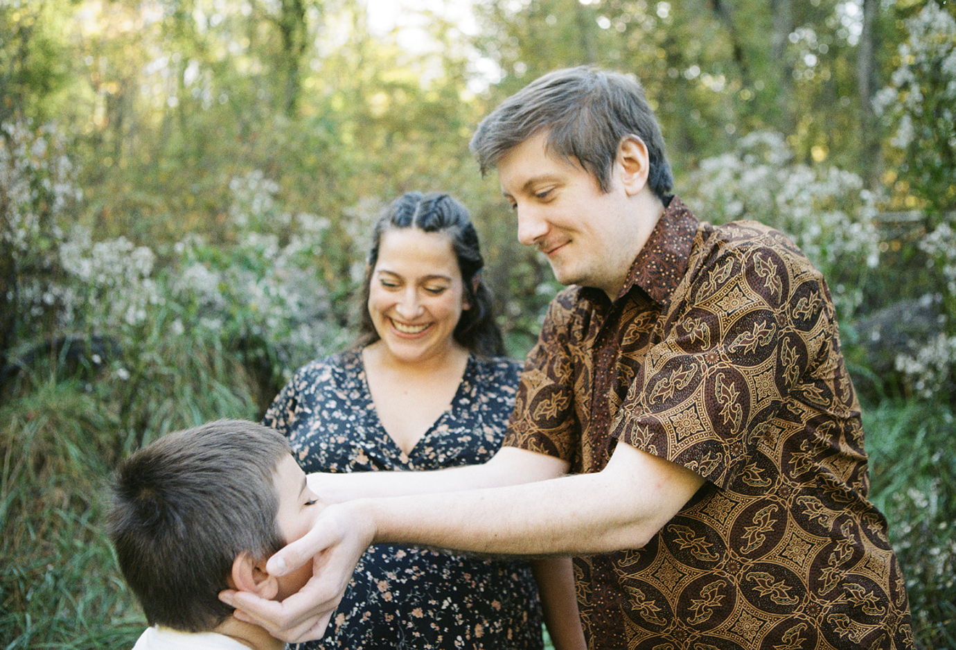 A family of three enjoying each other's company outdoors, with lush green foliage in the background. The father is playfully holding the son's face while the mother looks on smiling.