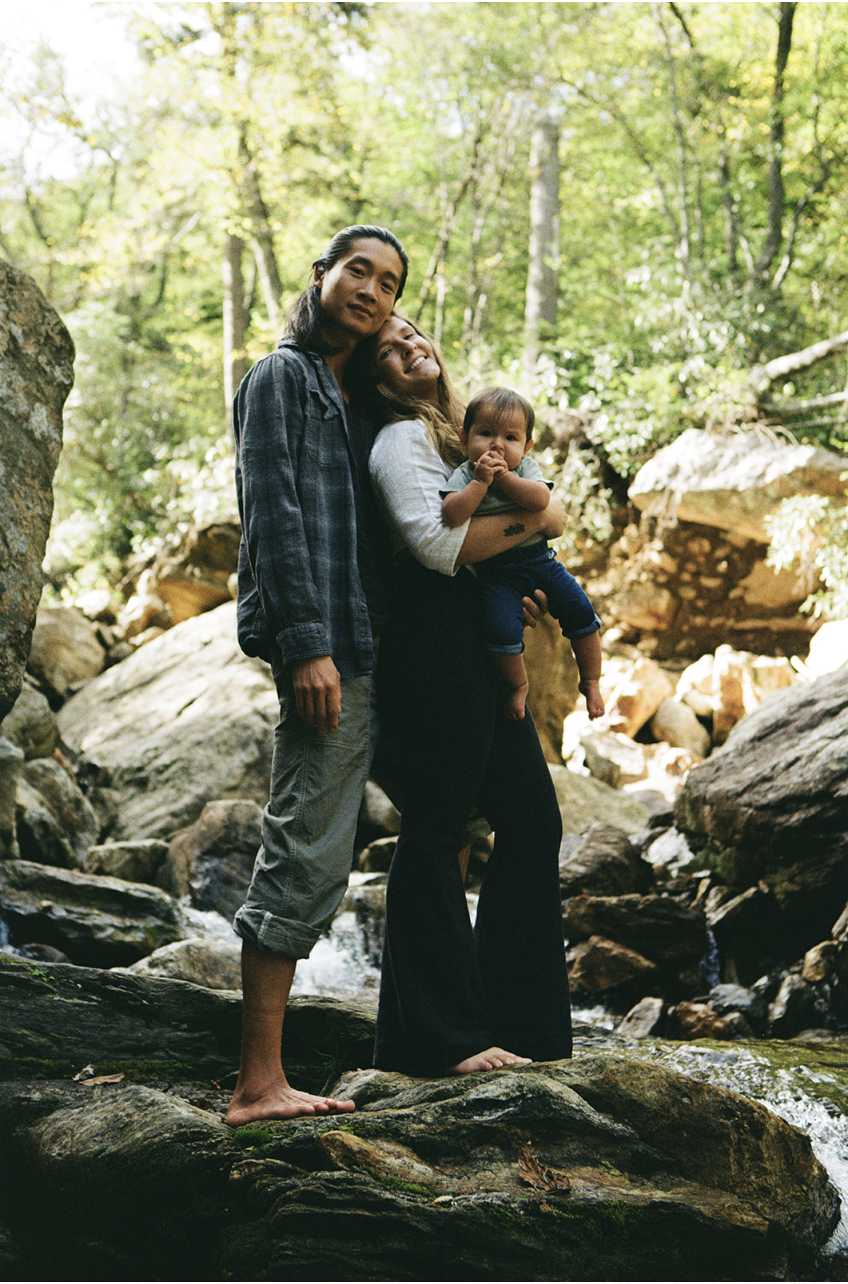 A family of three standing barefoot on rocks in a forest stream. The mother, holding a toddler, is smiling; the toddler is also smiling and suckling their finger. The father stands close, looking at the camera. Tall trees and rocks surround them, wit