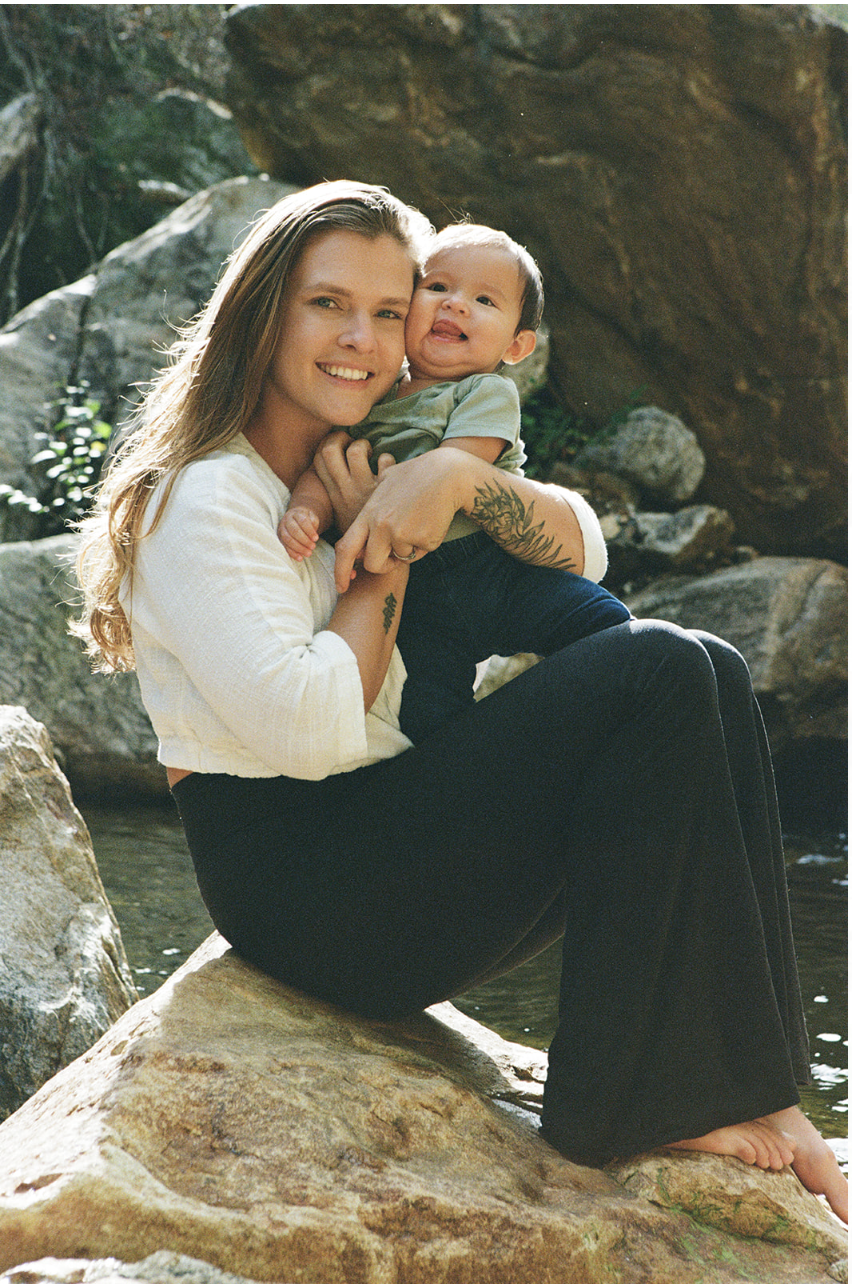 A woman sitting on a rock near water, holding a smiling baby in her arms.