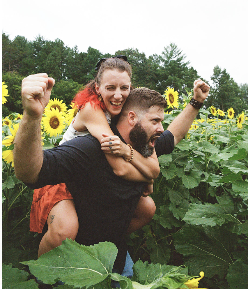 A couple enjoying a sunny day in a sunflower field, with the woman on the man's back, both fists raised in a gesture of excitement and happiness.