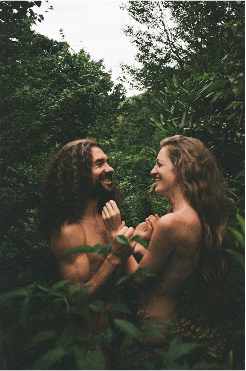 A man and woman are smiling and holding hands in a lush green outdoor setting, surrounded by dense foliage.