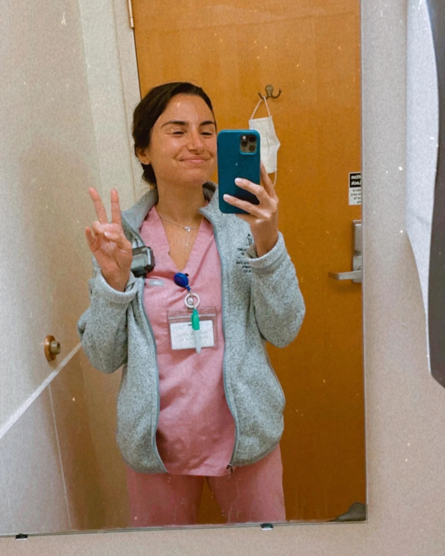 A young woman in medical scrubs taking a selfie in a hospital room mirror, making a peace sign with her right hand. She is wearing a gray zip-up jacket over pink scrubs, has a hospital ID badge around her neck, and a phone holder attached to her jacket.