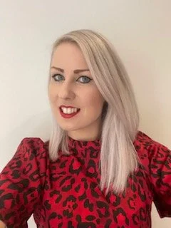 A woman with platinum blonde hair wearing a red and black leopard print top, smiling at the camera.