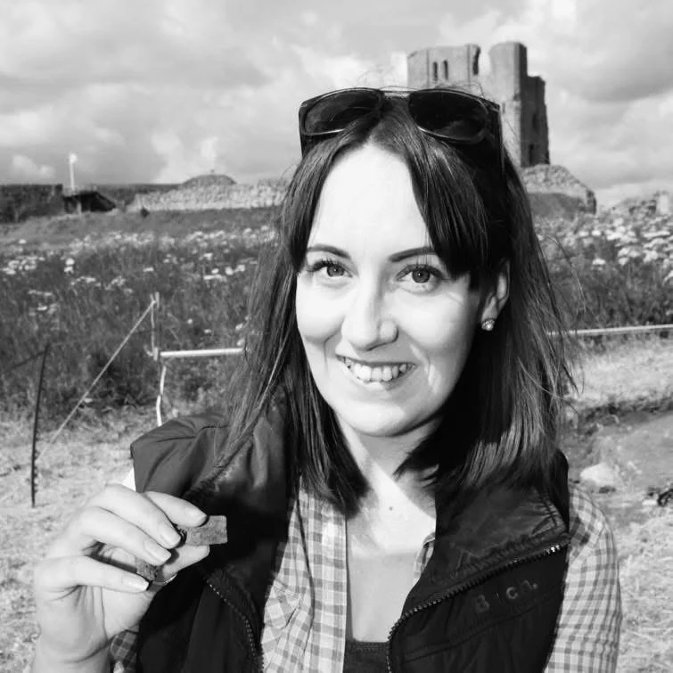 A woman with shoulder-length dark hair and sunglasses on her head, smiling and wearing a checkered shirt and vest, outdoors with ruins and cloudy sky in the background.