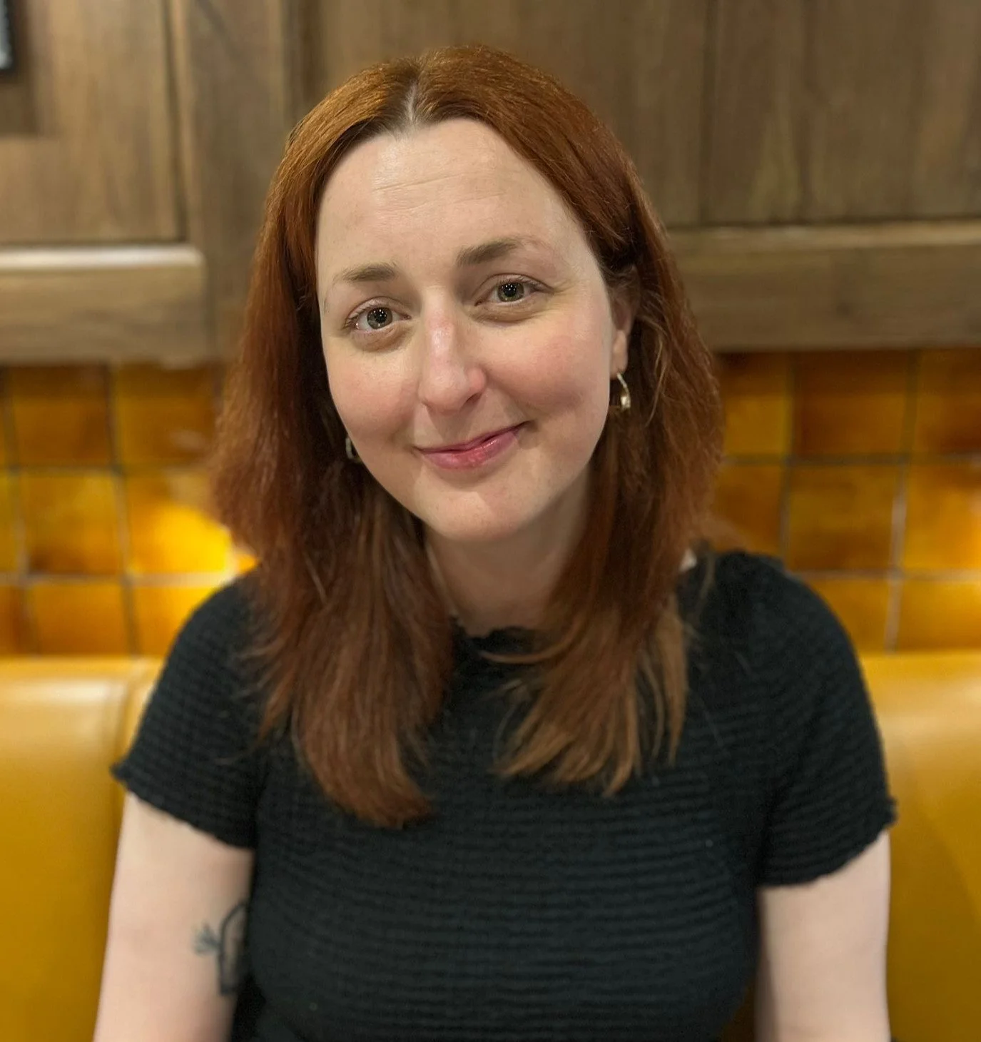 A woman with shoulder-length red hair, wearing a black top, smiling in a restaurant with wood-paneled walls and yellow upholstered seating.