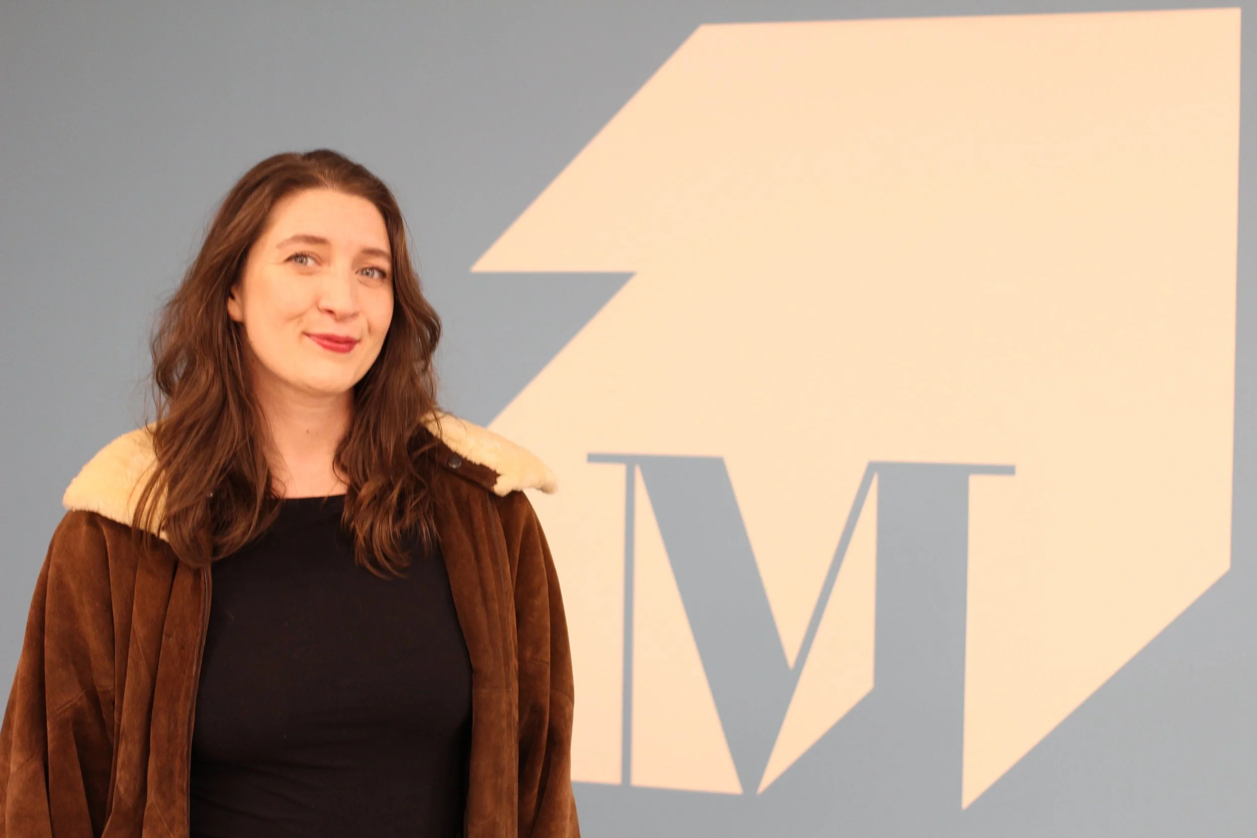 A woman with long brown hair wearing a brown jacket with a cream collar, standing in front of a large blue wall with a light-colored abstract logo and the letter ‘M’
