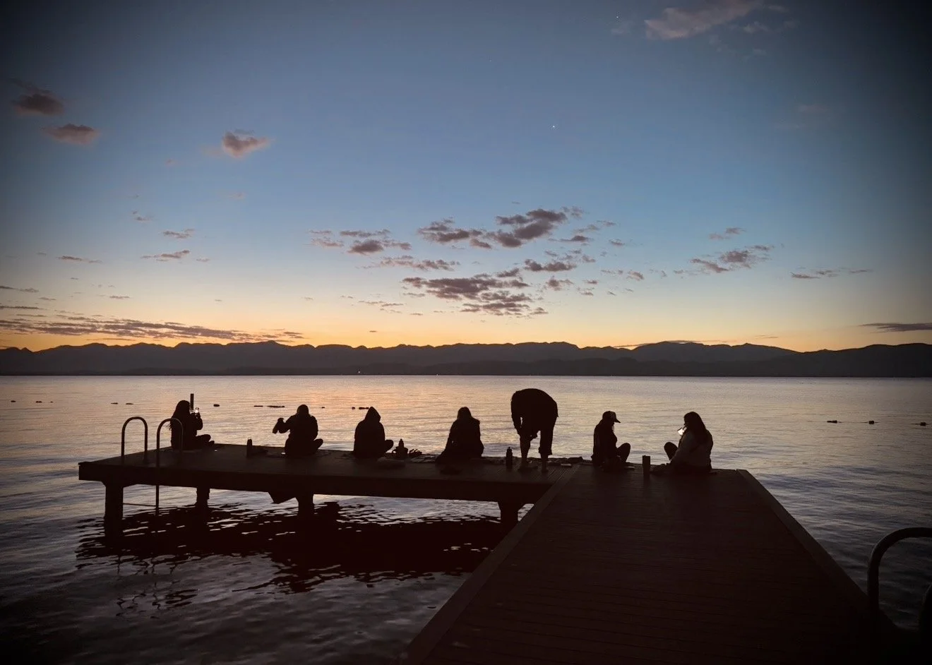Silhouettes of people sitting and standing on a wooden dock at sunset over a calm lake, with mountains in the background and a mostly clear sky with some clouds.