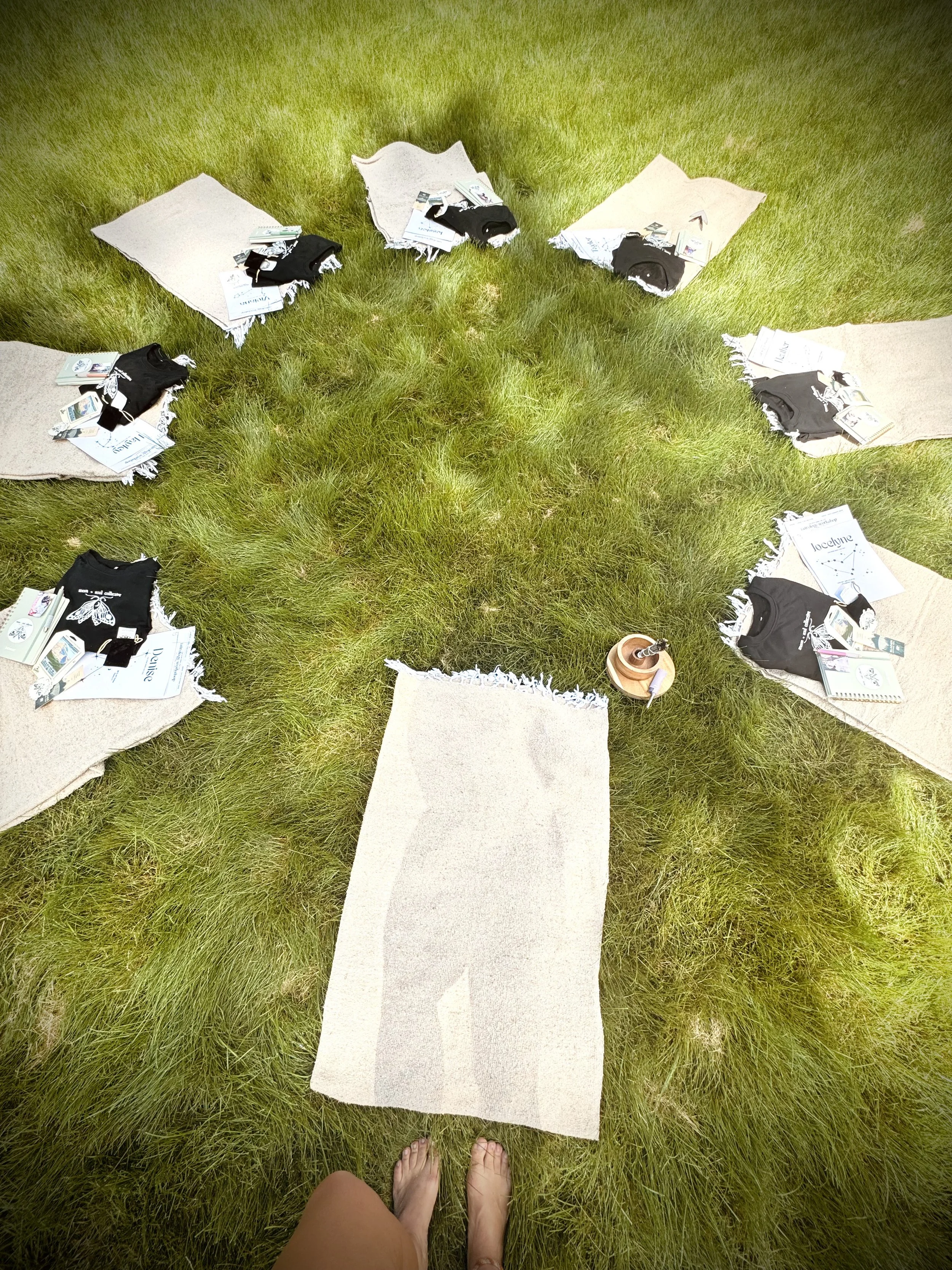 Outdoor scene with a person standing on a white rug on grass, surrounded by eight other rugs each with clothes and tags on them, and a small wooden bowl with a brush inside.