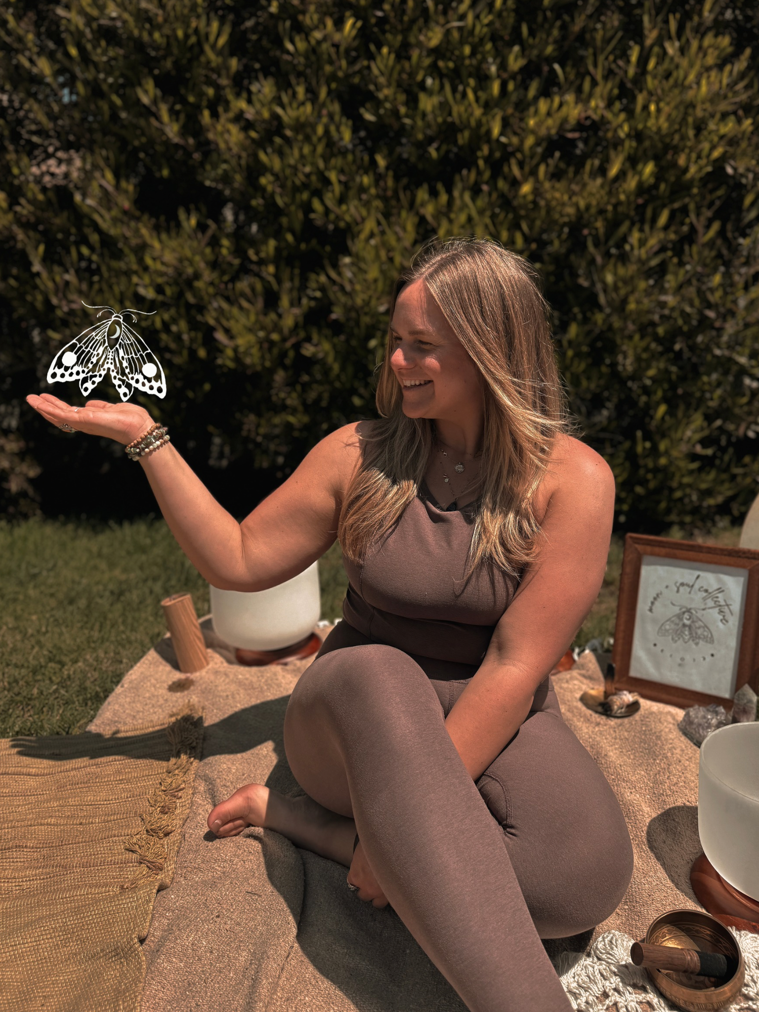 A woman sitting outdoors on a mat, holding her knee with one hand and extending her other hand, from which a digitally added butterfly is flying. She is smiling and surrounded by decorative objects and framed art.