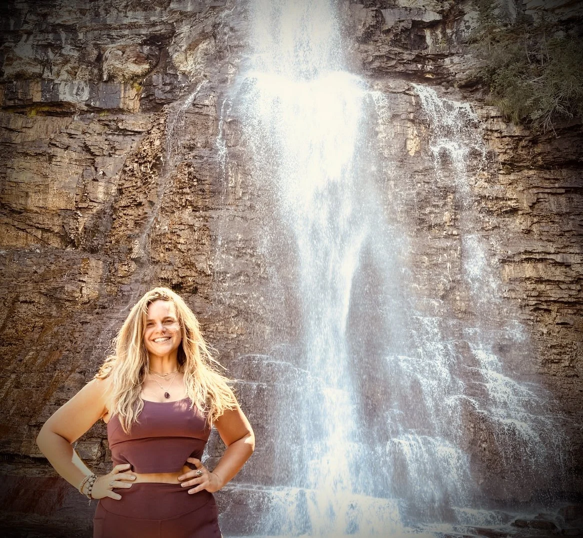 A woman with long blonde hair wearing a dark purple sleeveless top and matching skirt, smiling and standing with hands on hips in front of a tall waterfall cascading down a rocky cliff.