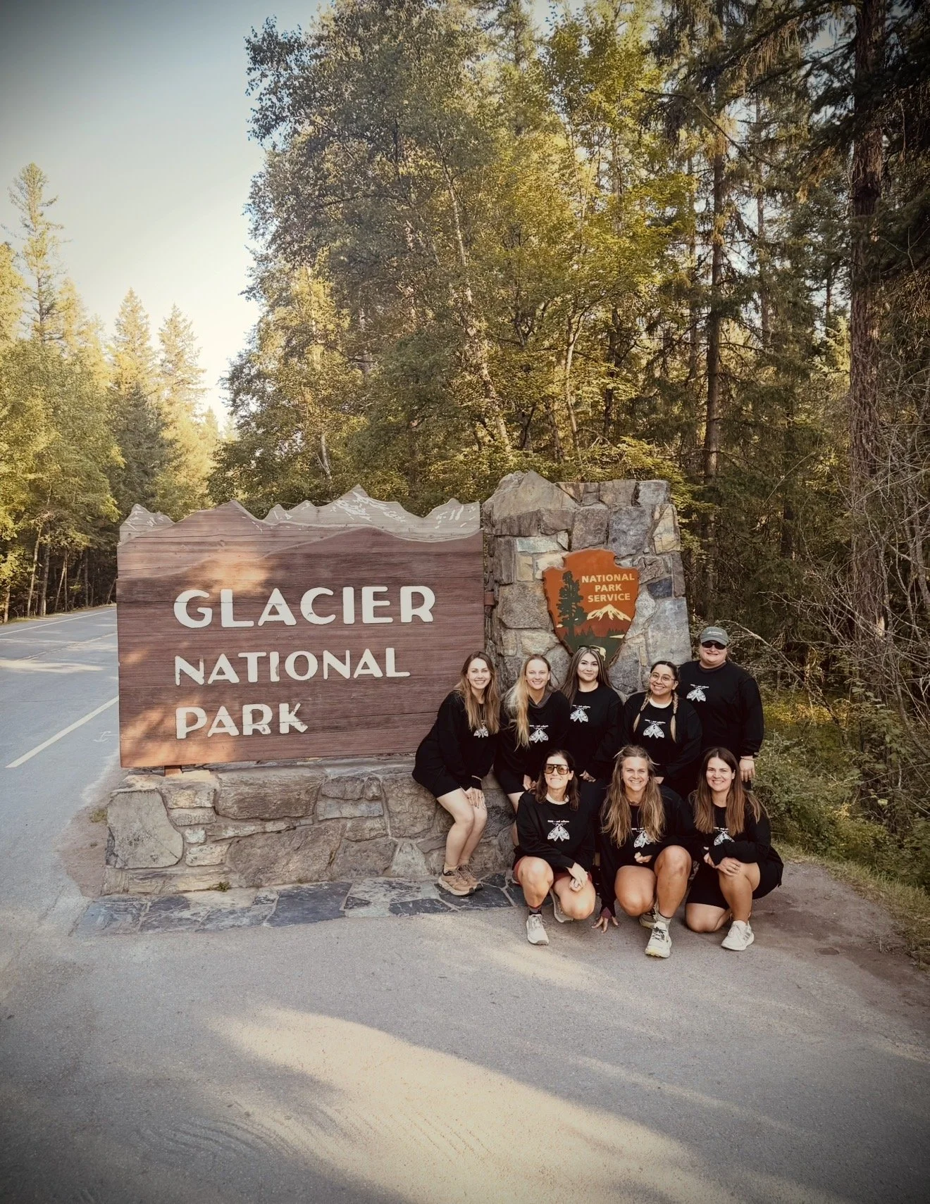 A group of nine people posing in front of the Glacier National Park sign, surrounded by trees and forest, during daytime.