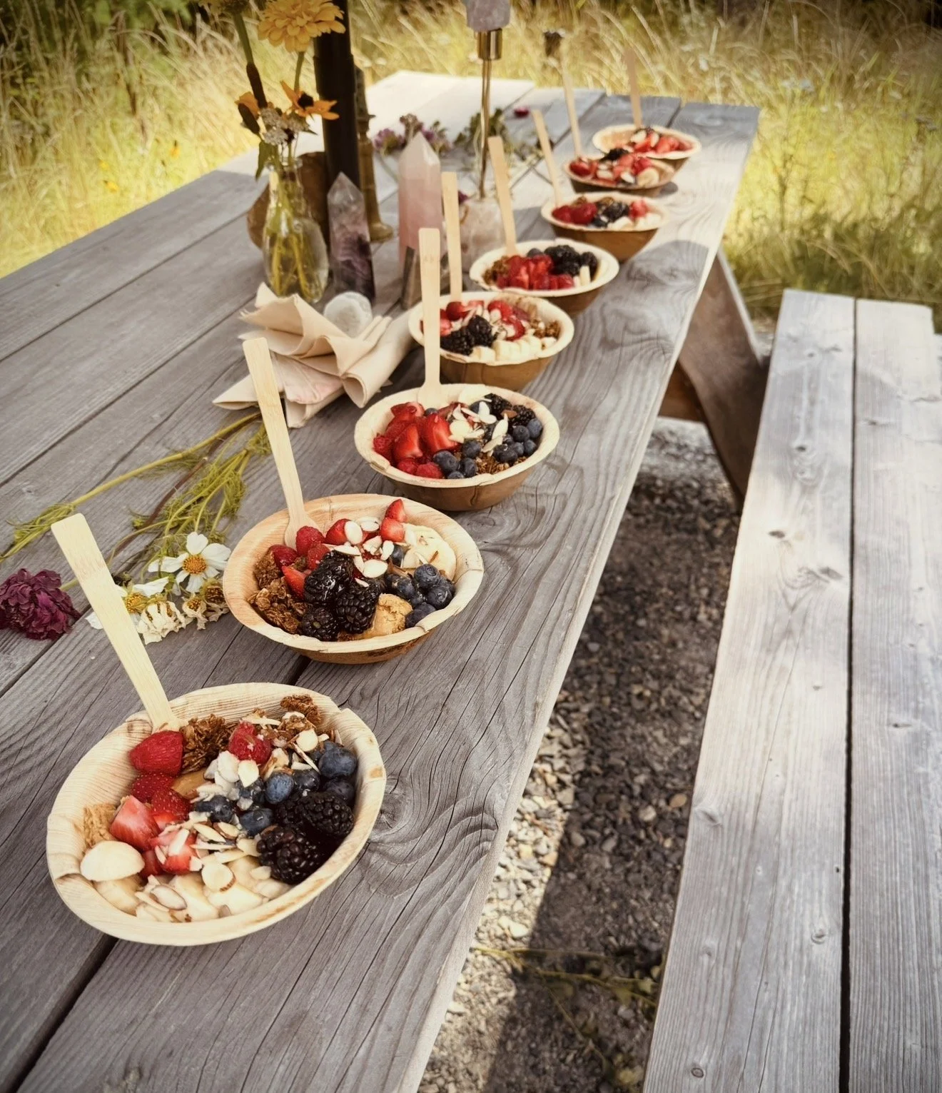A rustic outdoor scene with a long wooden table displaying bowls of berry-topped desserts, garnished with sliced almonds and fresh berries, set against a grassy background with decorative flowers and candles.