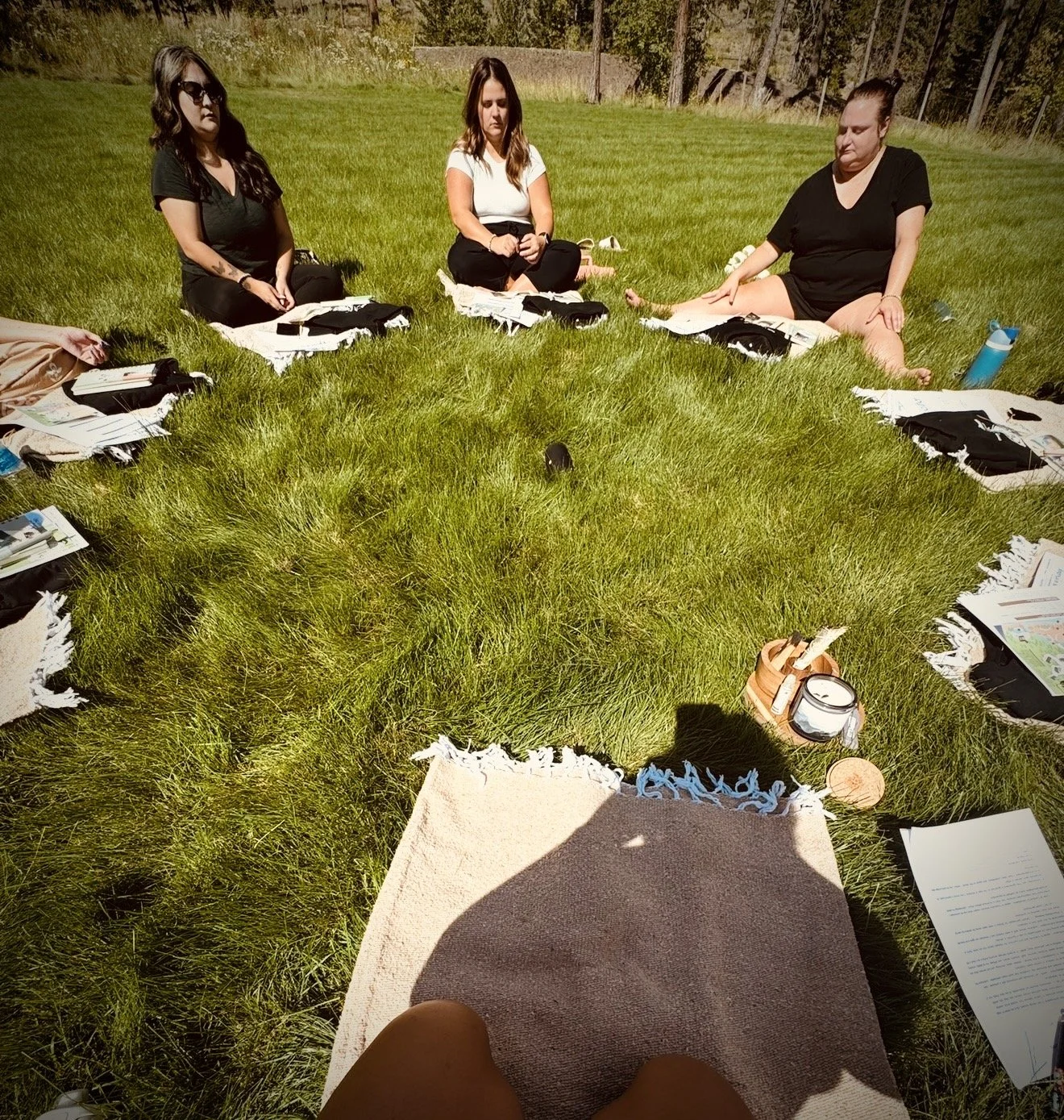 Three women seated on grass in a circle with various papers, bags, and drinks around them on a sunny day.