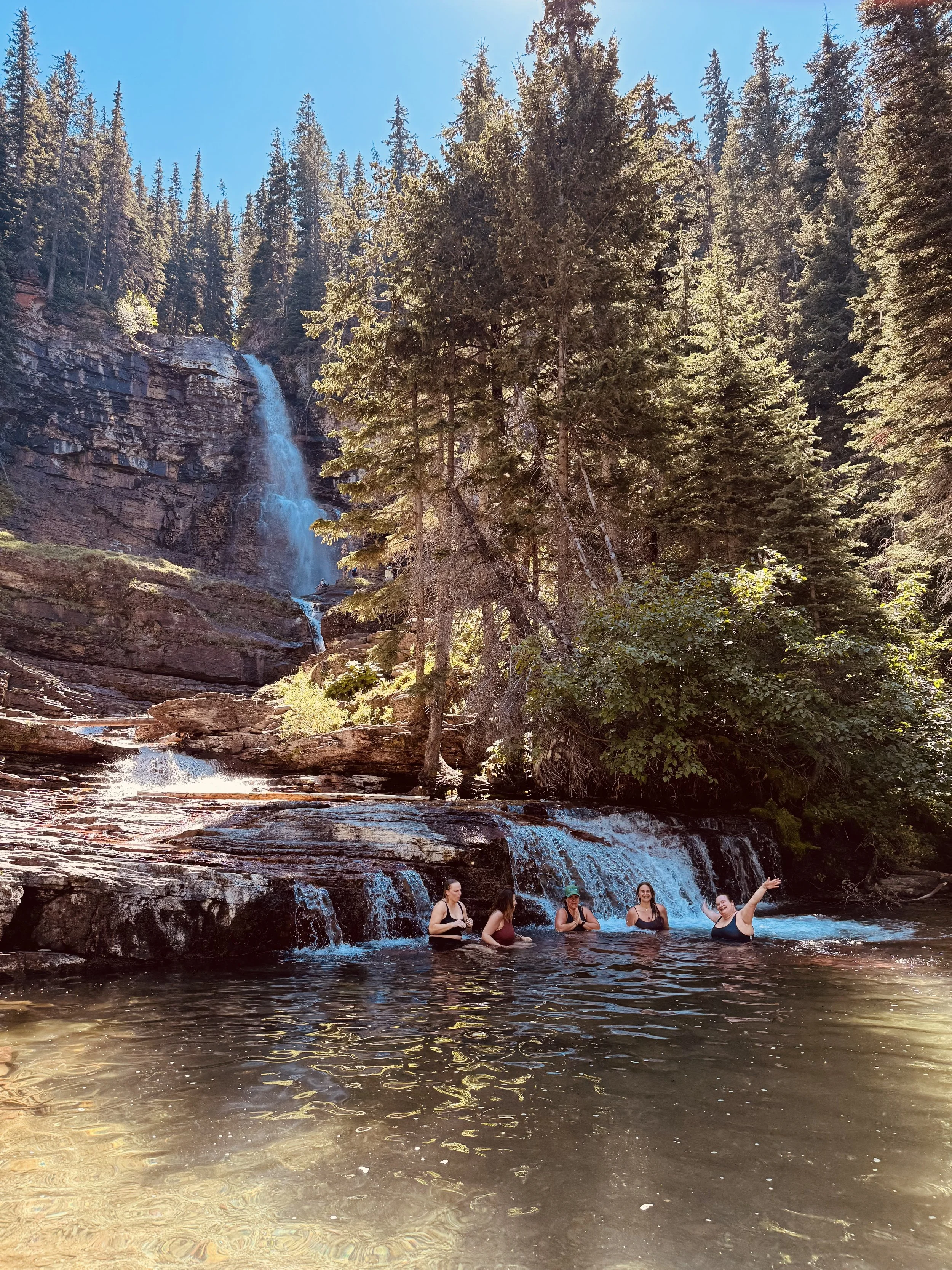 Four people swimming and standing in a natural pond at the base of a waterfall in a forested mountain area.