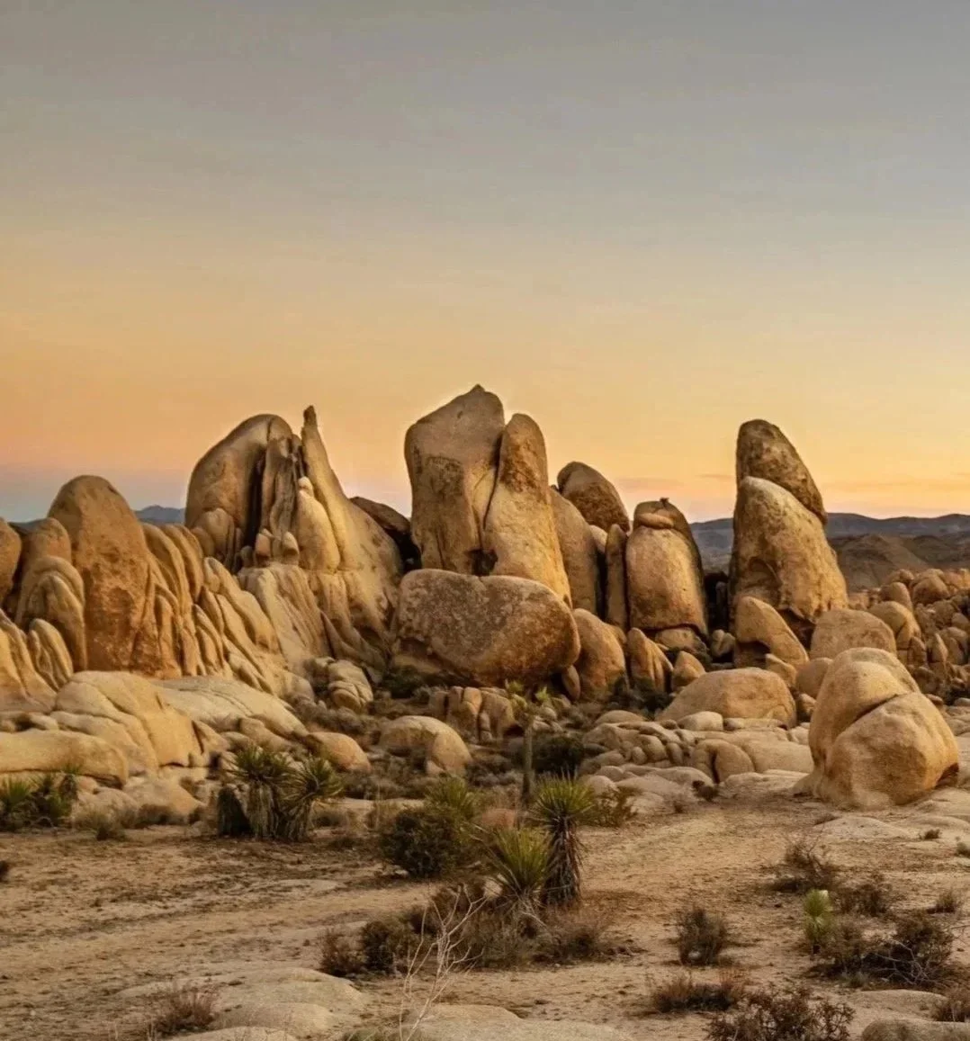 A desert landscape with large rock formations at sunset, sparse desert vegetation, and a colorful sky with shades of yellow, orange, and blue.