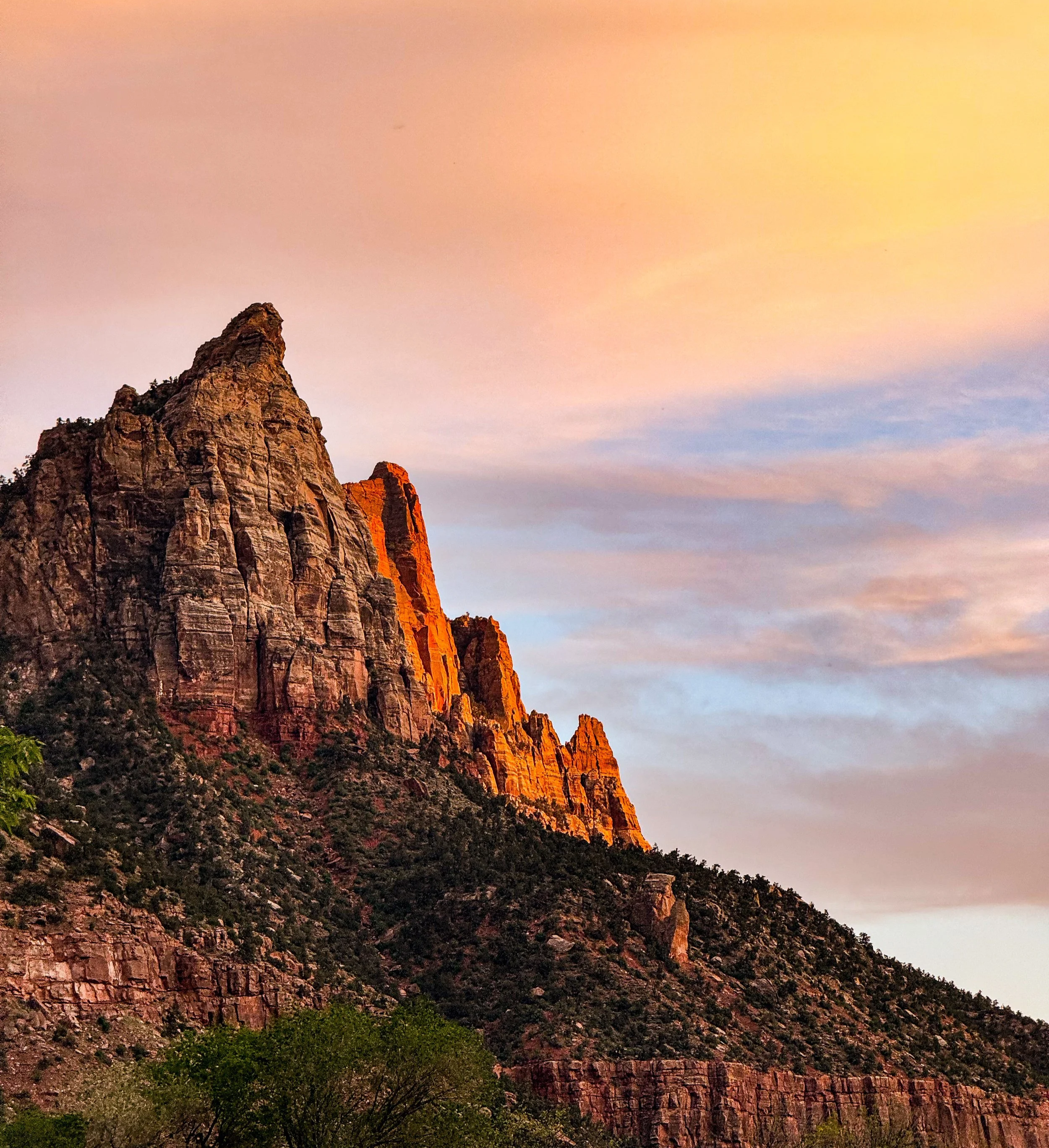Sunset over rocky mountain peaks with orange light illuminating the tops, partly cloudy sky, green trees at the base.