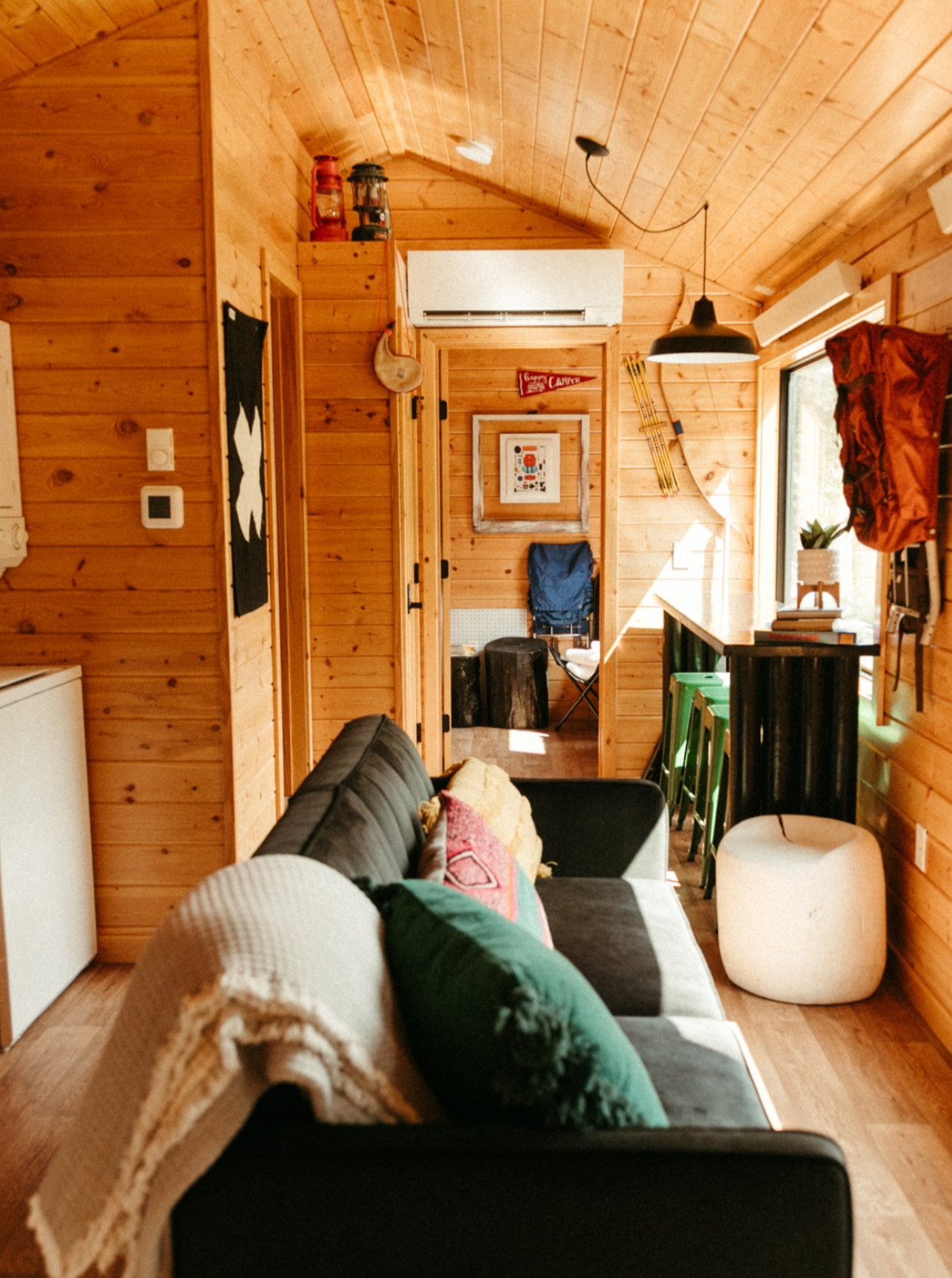 Cozy living room with wooden walls, a black sofa with colorful pillows, large window with curtains, and a small kitchen bar area with stools. Decor includes lanterns, framed pictures, and camp-themed signs.