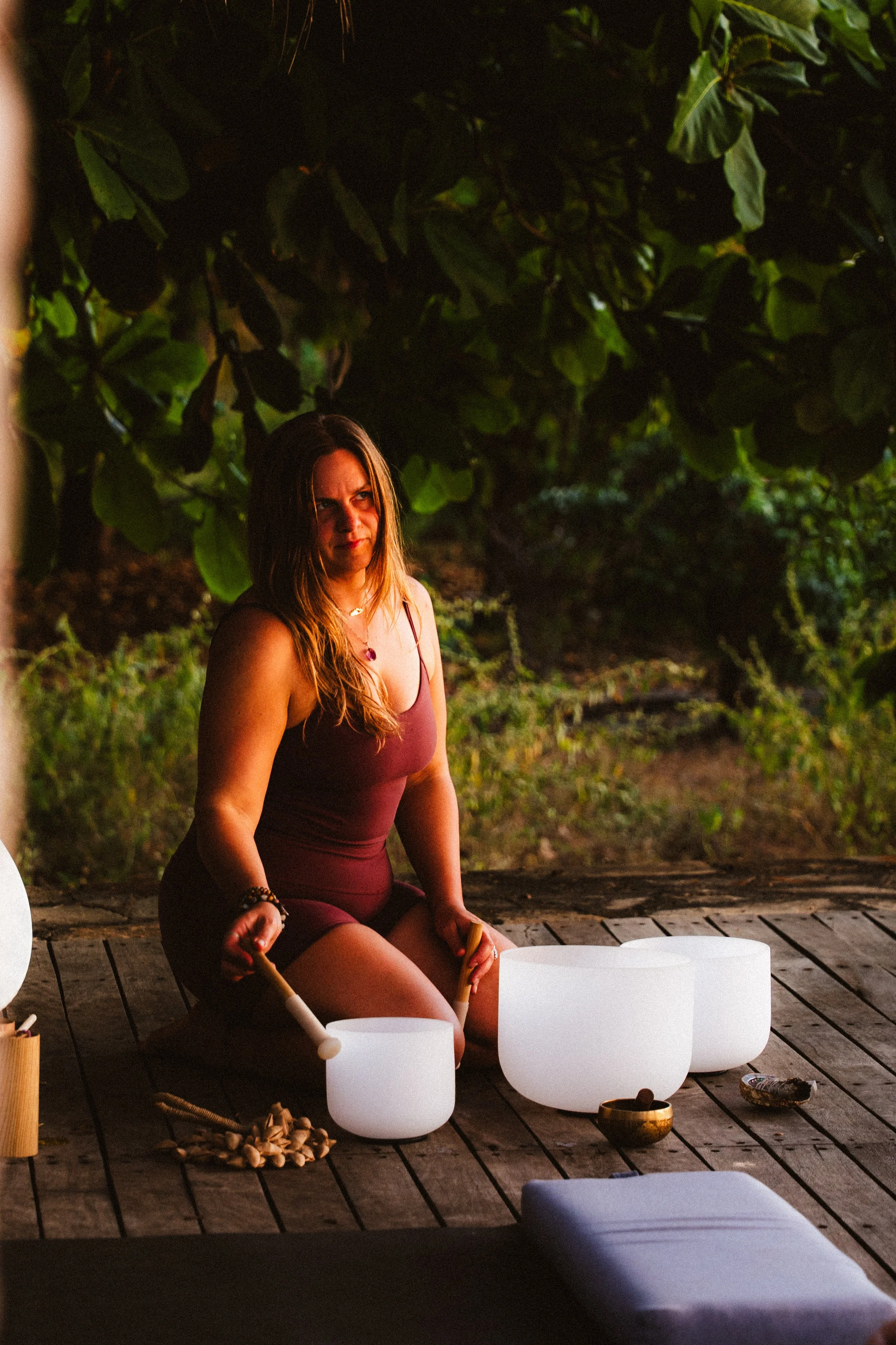 A woman in a maroon dress is sitting on a wooden deck, holding two singing bowls, with another singing bowl and a mallet nearby, surrounded by lush green foliage at dusk.