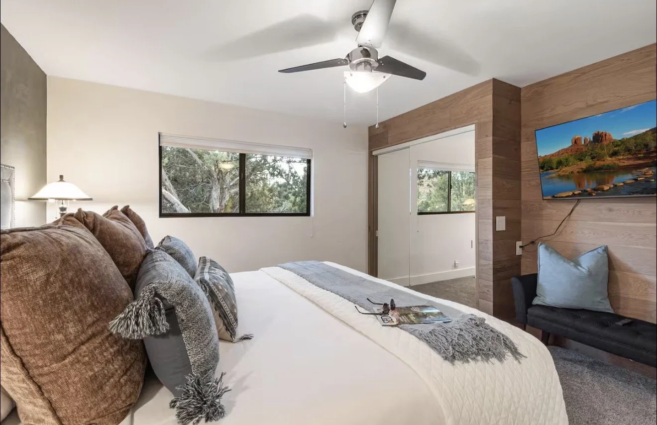 A bedroom with a large bed, white bedding, and decorative pillows, flanked by nightstands with lamps, featuring a wooden ceiling fan, two abstract paintings on the wall, a large window with patterned curtains, and a striped rug on the floor.