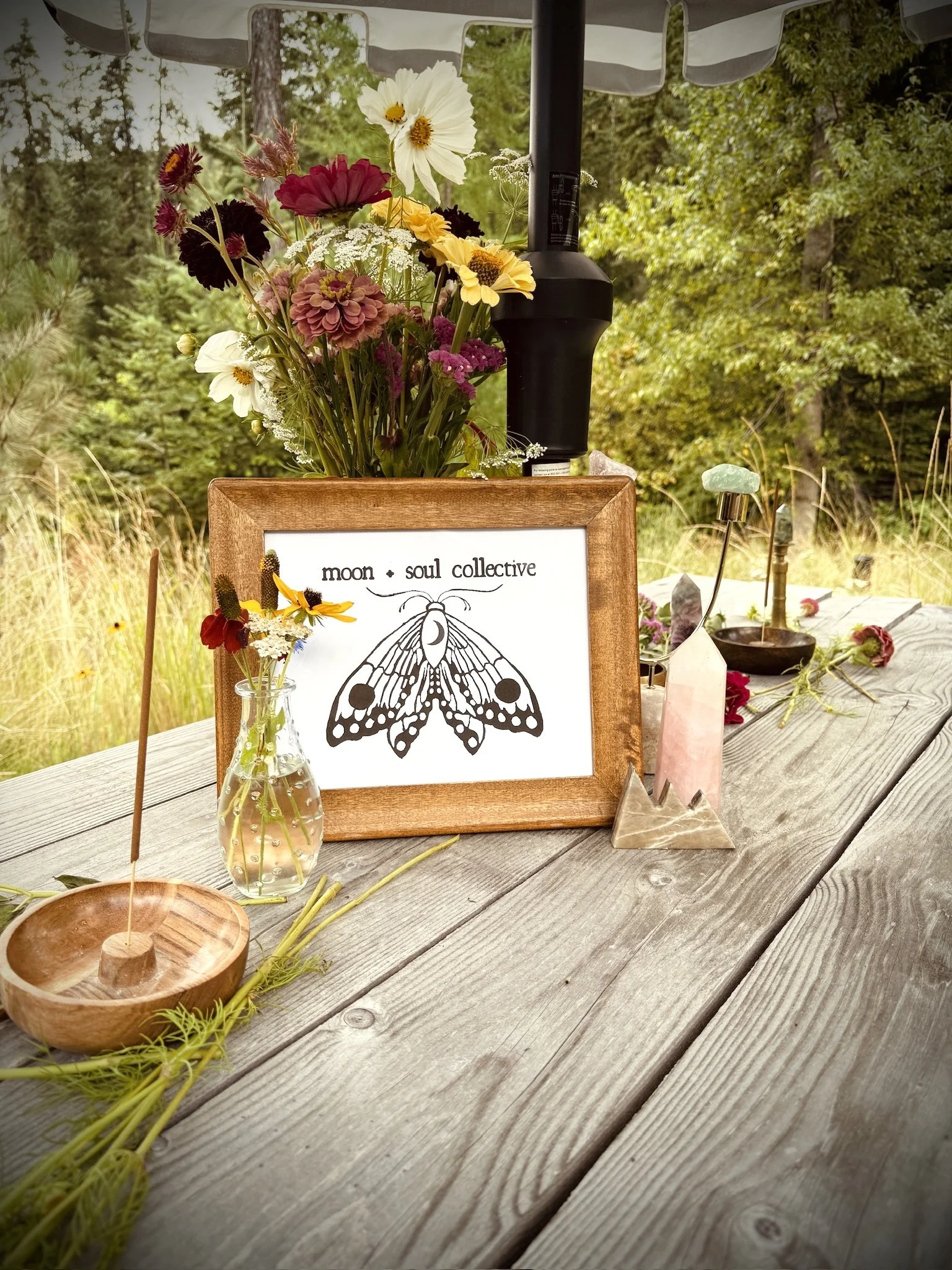 A table outdoors with a centerpiece of a framed illustration of a moth labeled "moon + soul collective," surrounded by flowers, crystals, and decorative items under a shade umbrella amid trees and greenery.