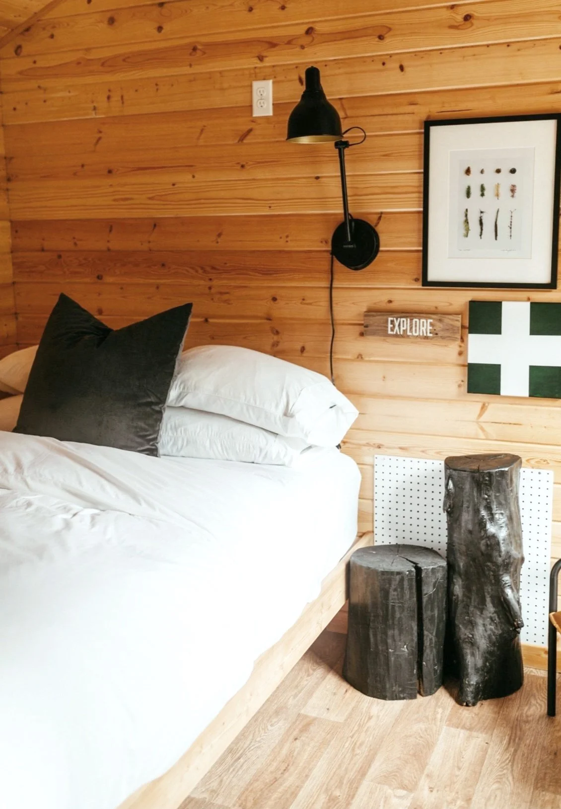 A cozy bedroom corner with a wooden wall, a bed with white linens and black pillows, wall-mounted black reading light, framed artwork, and wooden logs used as decor.