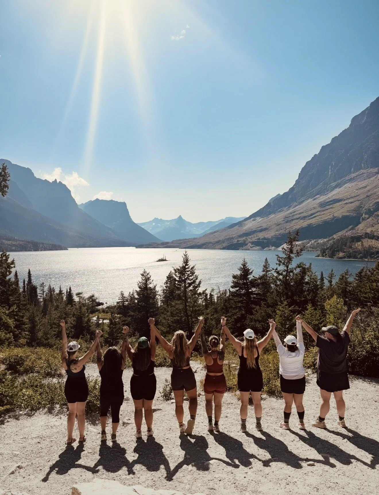 Nine people holding hands and raising their arms, standing on a trail overlooking a lake and mountains.