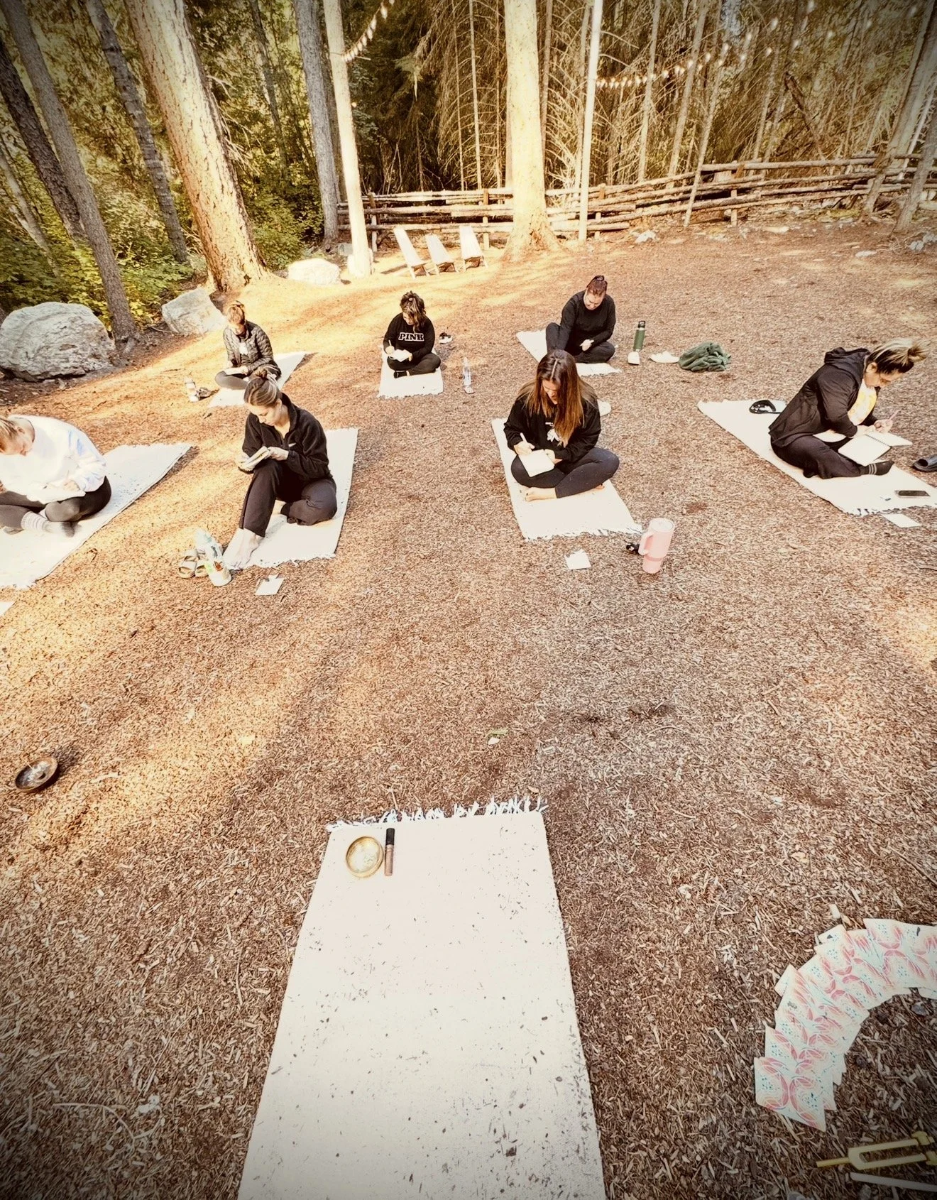 Women practicing yoga outdoors on mats in a wooded area, with some reading and drinking water.