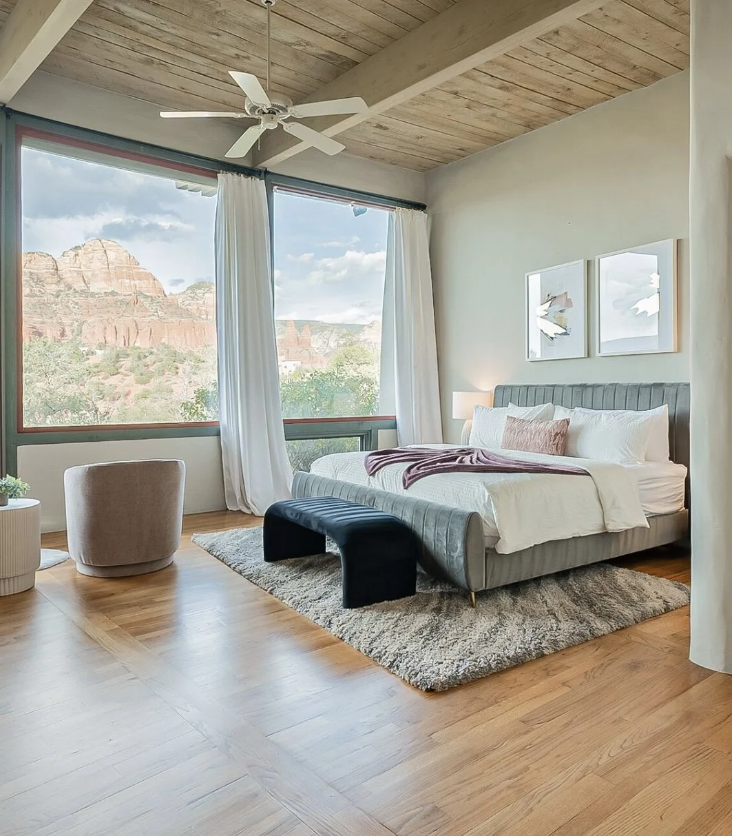 A bedroom with a large window showing red rock formations outside, a bed with white bedding and pillows, a black bench at the foot of the bed, a beige chair, a textured rug, and two framed artworks on the wall.