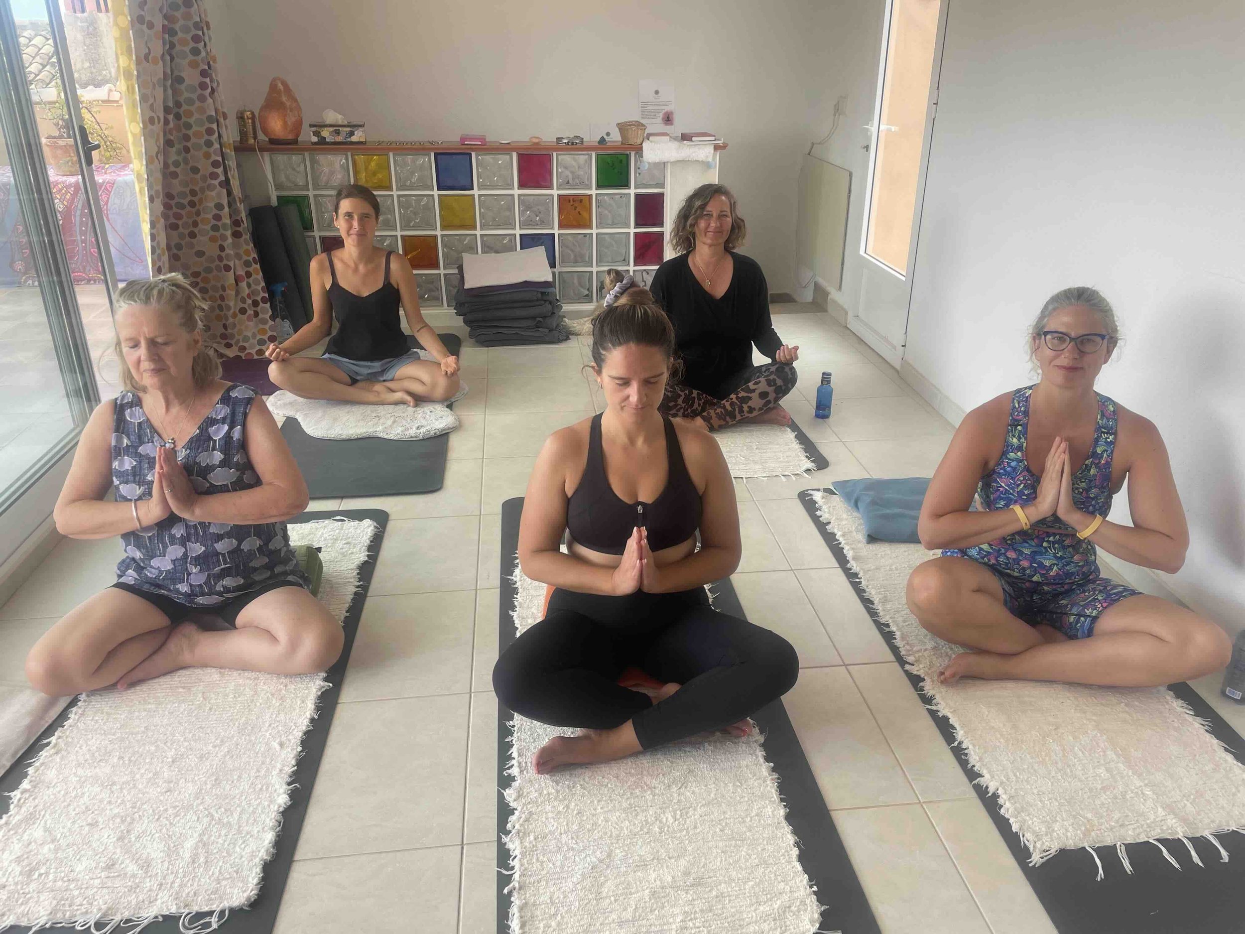 Group of people sitting cross-legged on yoga mats, practicing meditation indoors. They have their hands in a prayer pose. Background features shelves with colorful squares and a salt lamp on an Aum Retreat.