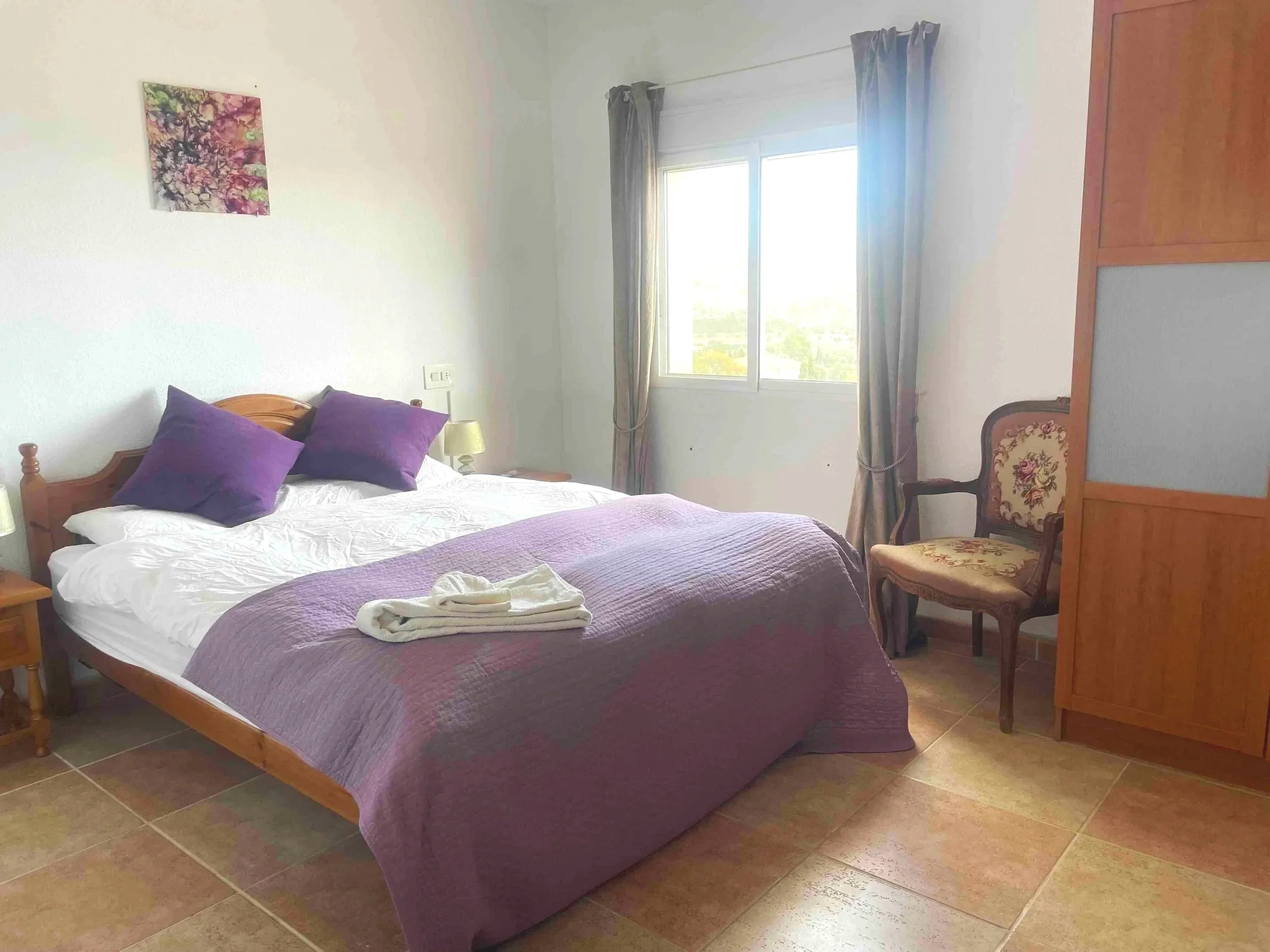 Casa Aum bedroom with a bed featuring purple pillows and blanket, wooden nightstand, window with curtains, upholstered chair, and artwork on the wall.