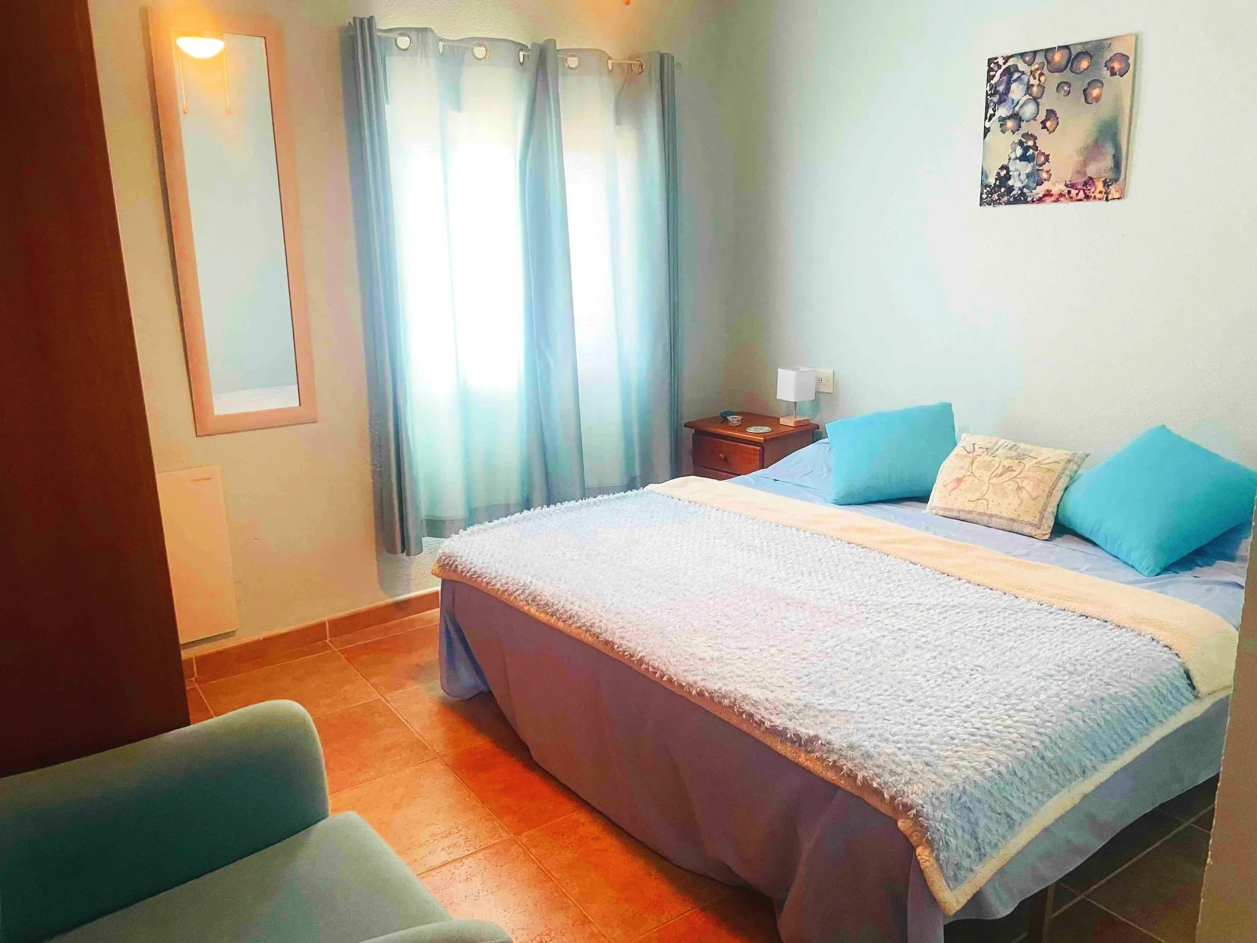 Casa Aum bedroom with a double bed dressed in light blue bedding, two teal pillows, and a decorative pillow, a wooden nightstand with a lamp, a wall-mounted mirror, curtains on a window, and a piece of abstract art on a light-colored wall.