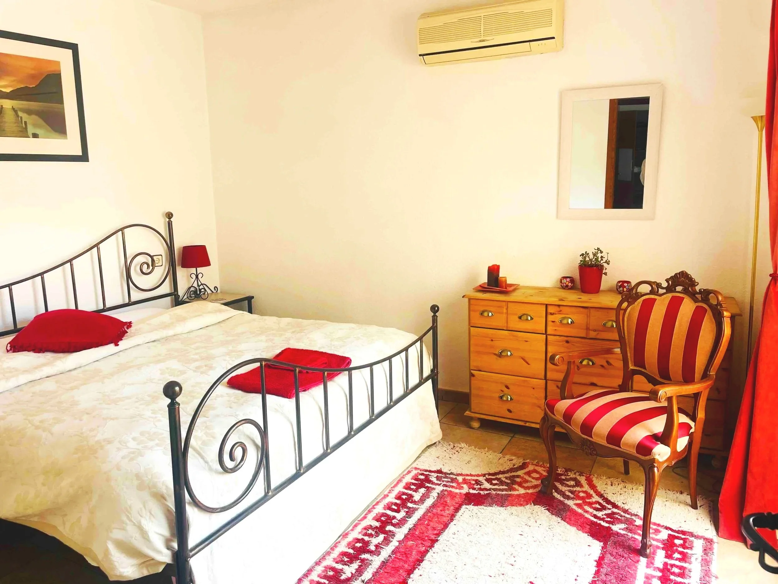 Casa Aum bedroom with a black metal bed frame, white bedding, and red accents, including a pillow and towels. There's a wooden dresser with a mirror above, a red and beige striped chair, and a red decorative rug.