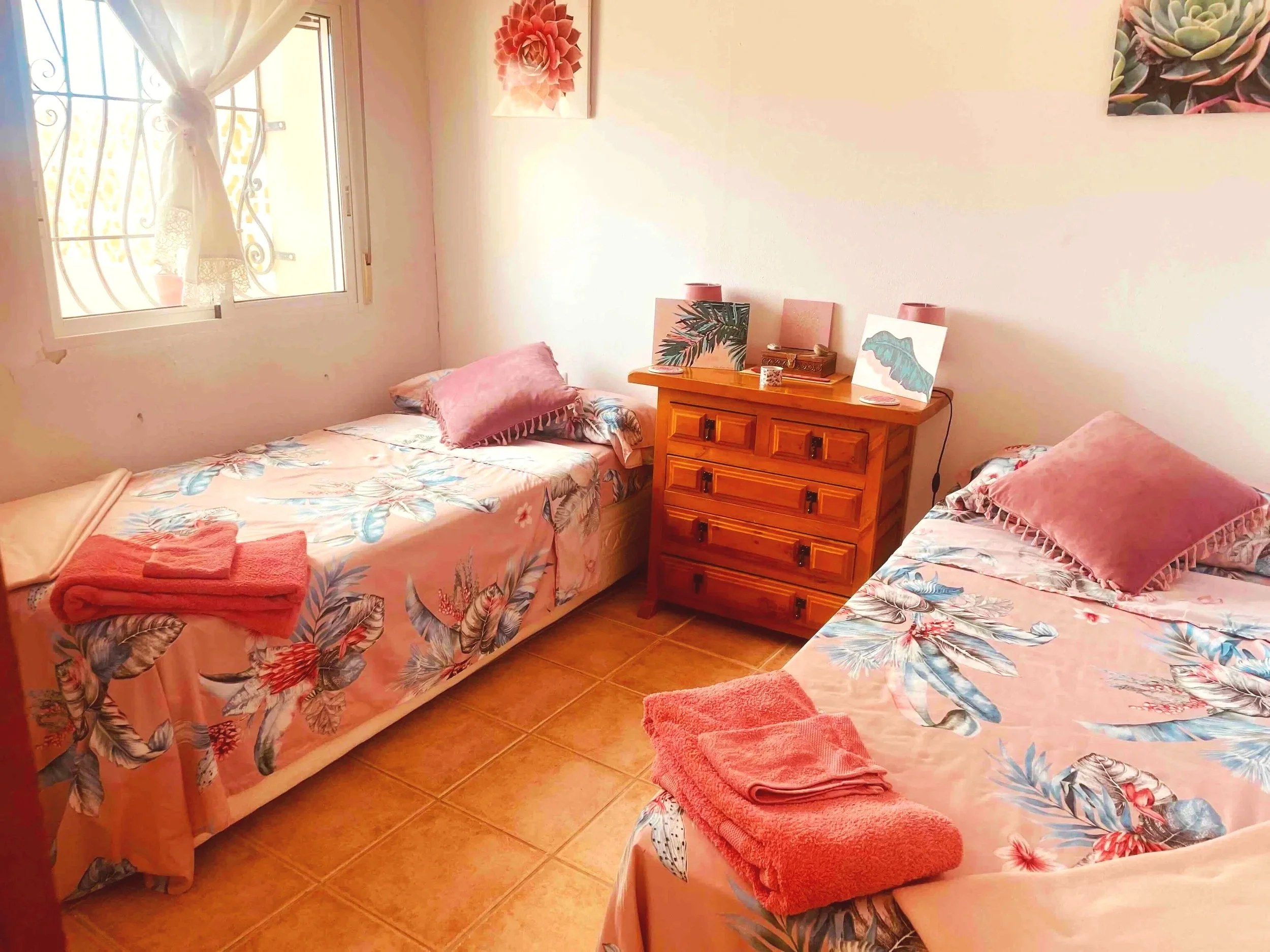 Casa Aum small bedroom with two twin beds featuring floral bedding, topped with pink pillows and neatly folded pink towels. A wooden dresser is positioned between the beds with decorative items and artwork above. 
