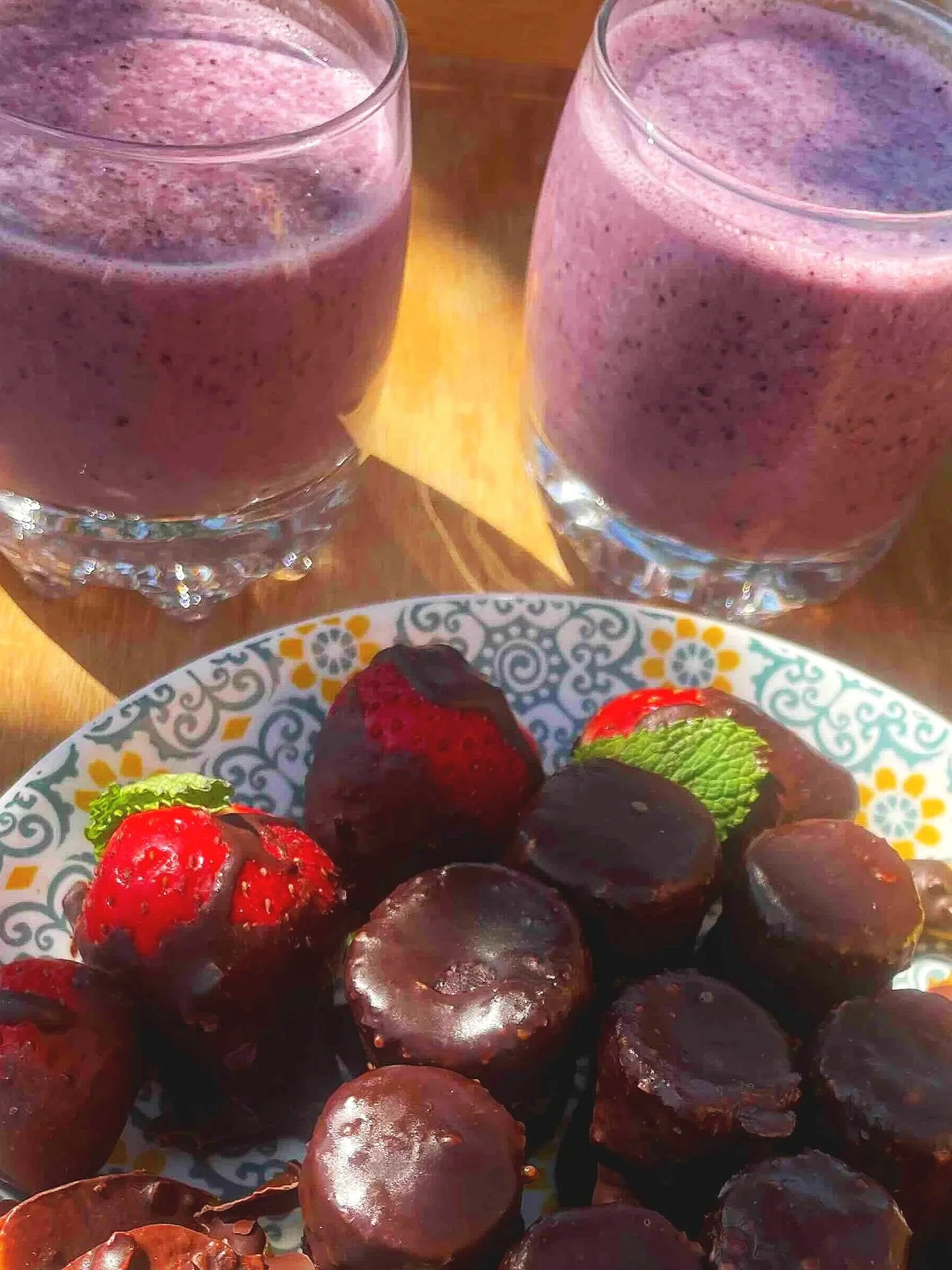 Two glasses of purple smoothies and a plate of chocolate-covered fruit including strawberries and peach slices on a wooden tray.