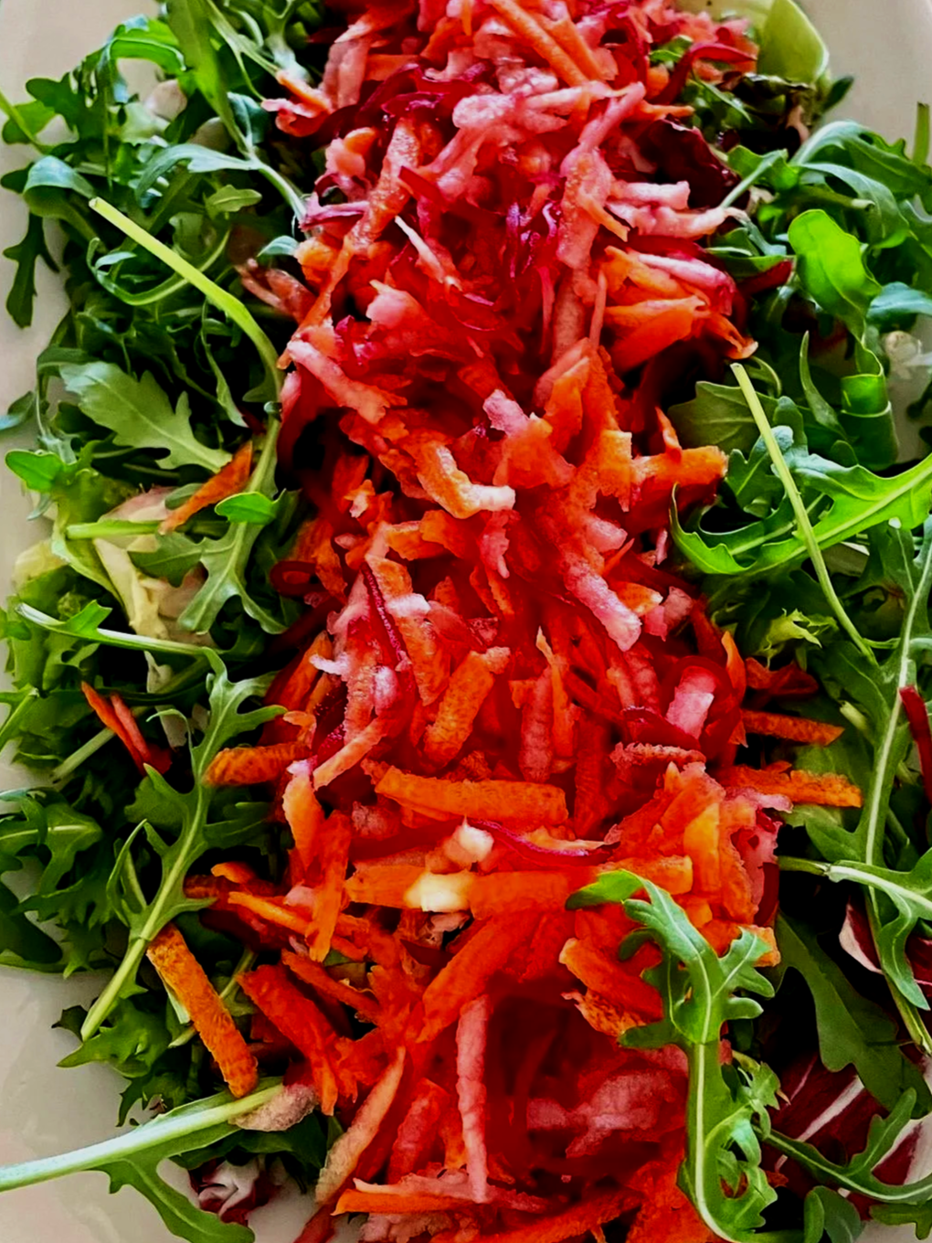 A mixed salad with shredded carrots, beets, and fresh arugula on a white rectangular plate, placed on a patterned tablecloth.