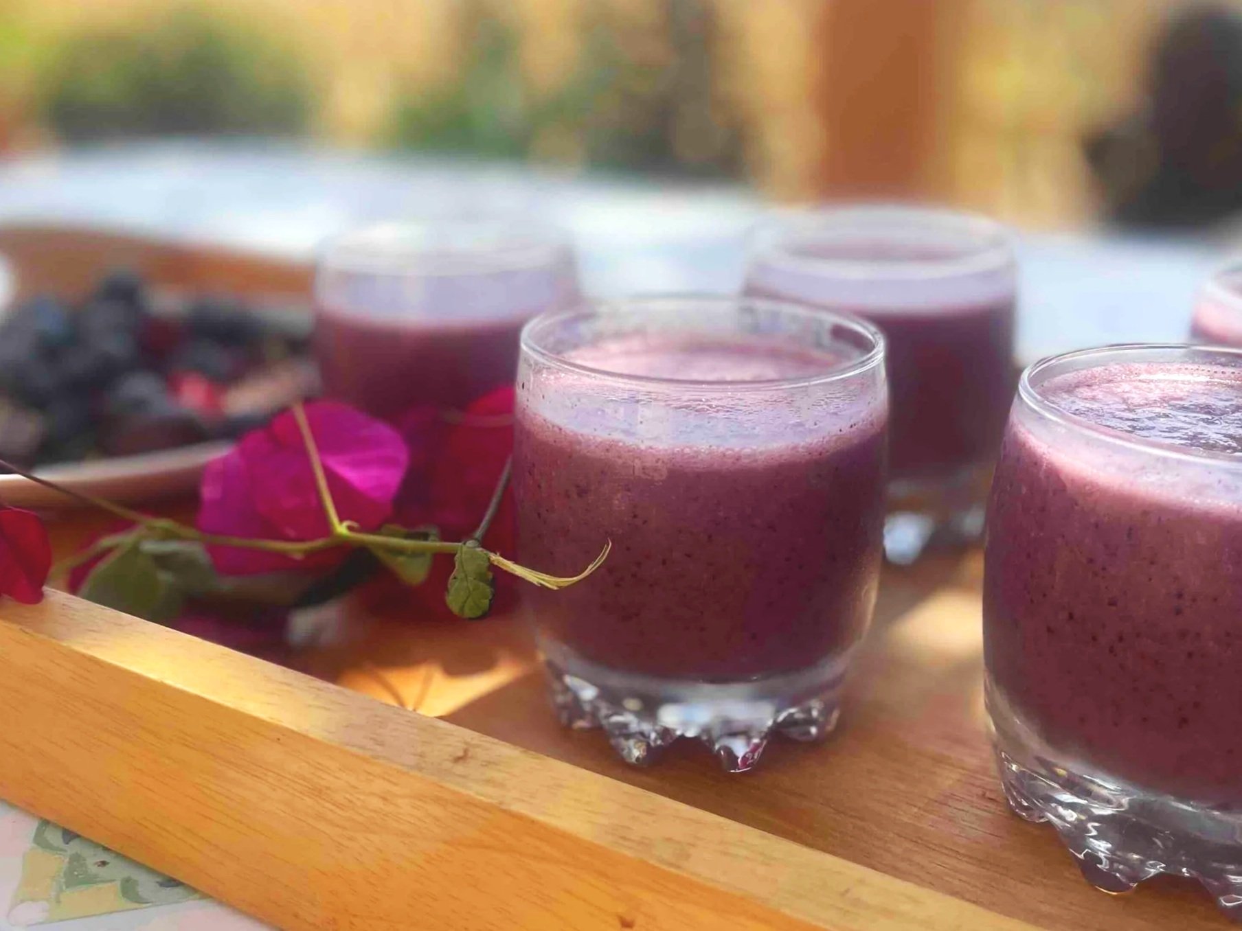 Small glasses filled with a purple smoothie on a wooden tray, accompanied by a plate of mixed berries and flowers for decoration.