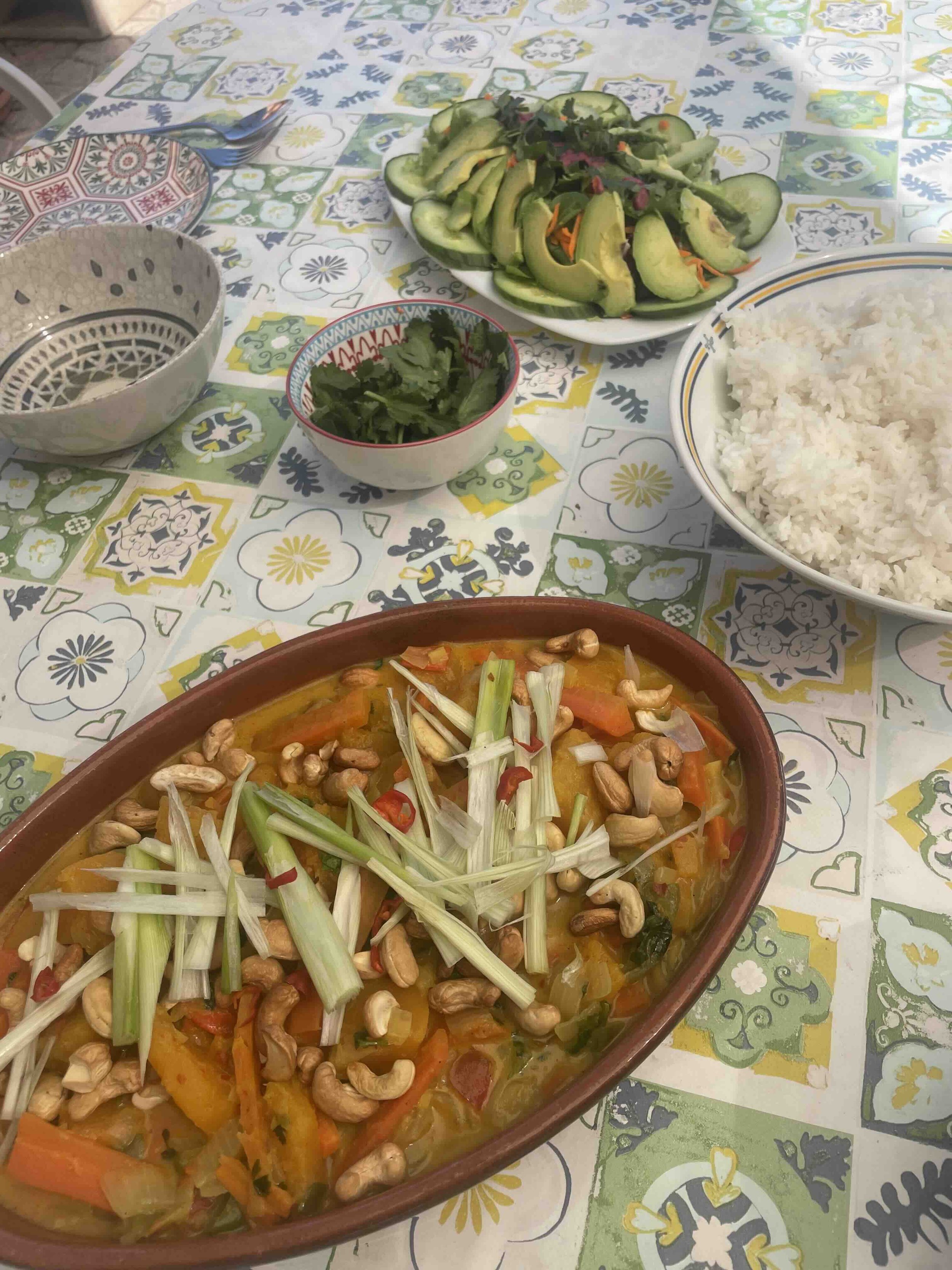 Colorful table setting with a bowl of vegetable curry garnished with cashews and leeks, plates of sliced avocado and cucumber salad, a bowl of white rice, and a small dish of herbs on a patterned tablecloth.