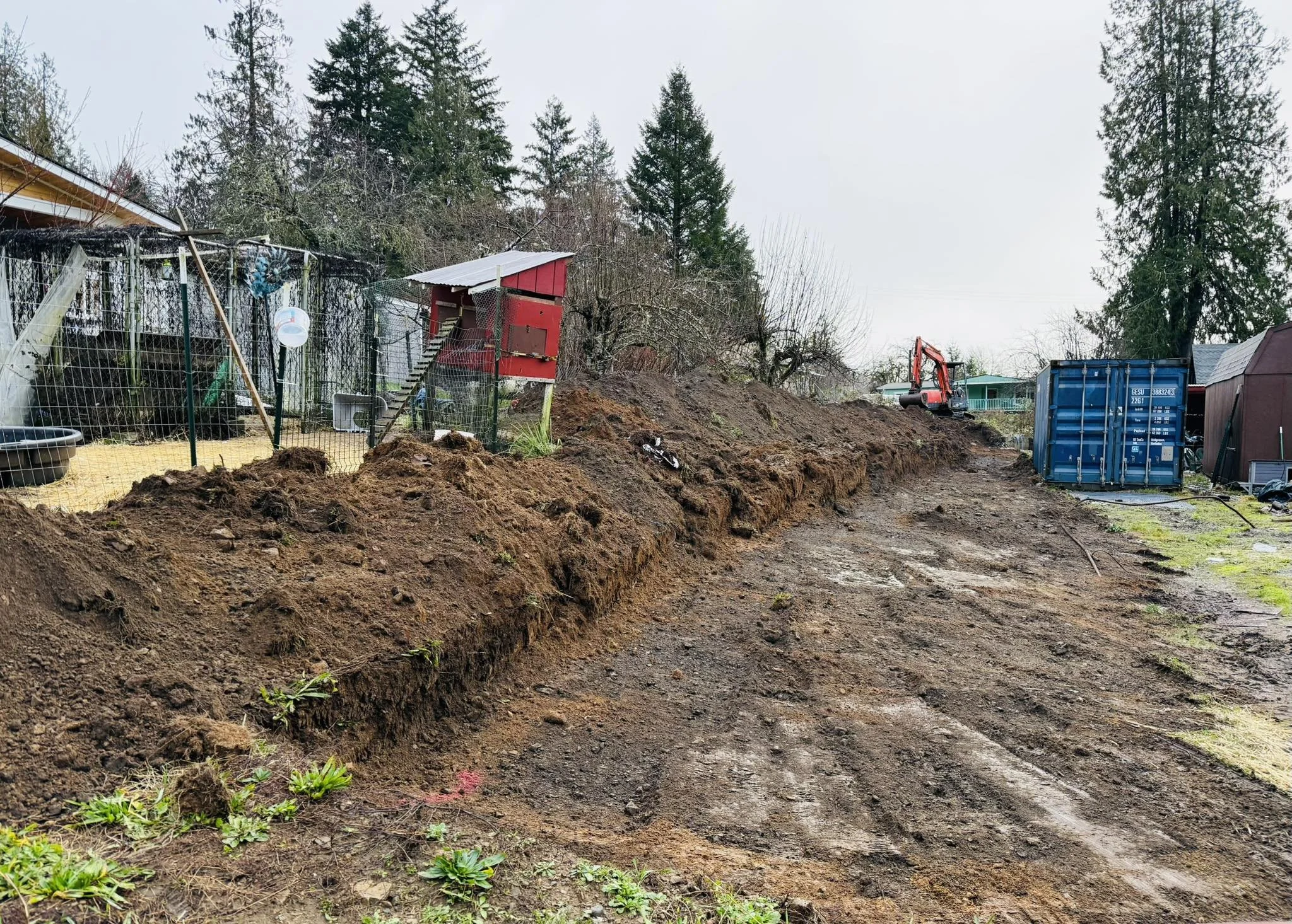 A construction site in a residential yard with soil excavation, a small red shed, construction equipment, a blue storage container, and trees in the background.