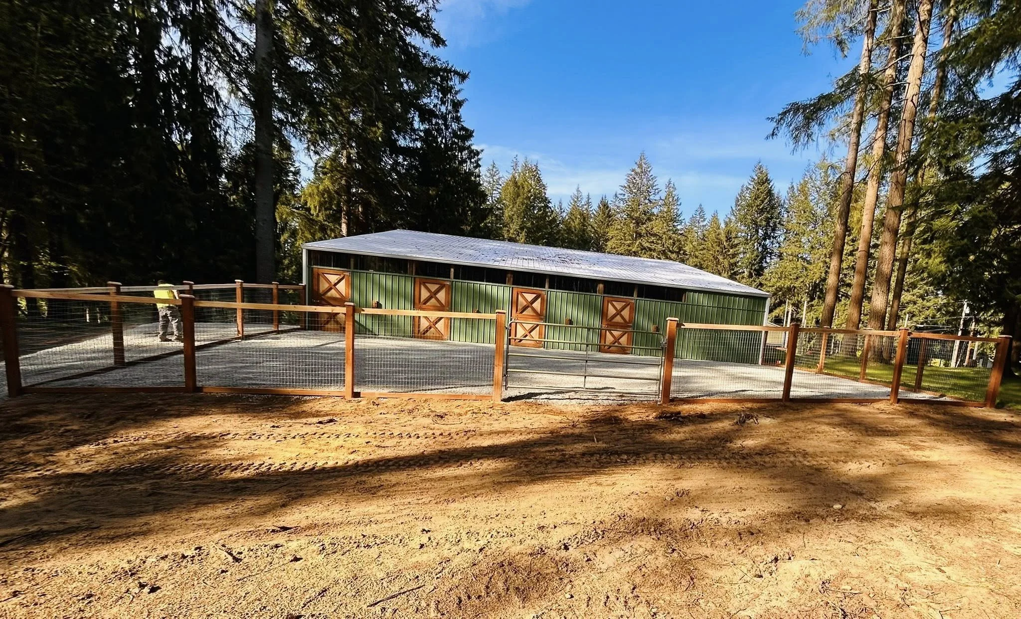 A green barn with brown doors surrounded by tall trees in a forested area, with a gravel area and wooden fence in the foreground, under a clear blue sky.