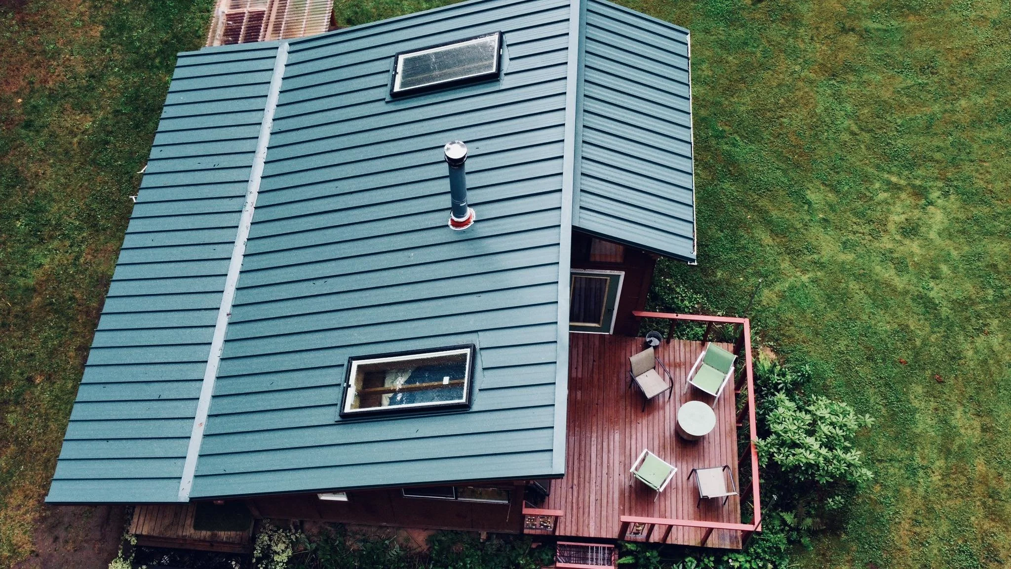Aerial view of a house with a blue metal roof and a wooden deck patio with outdoor chairs and a table, surrounded by green grassy yard.