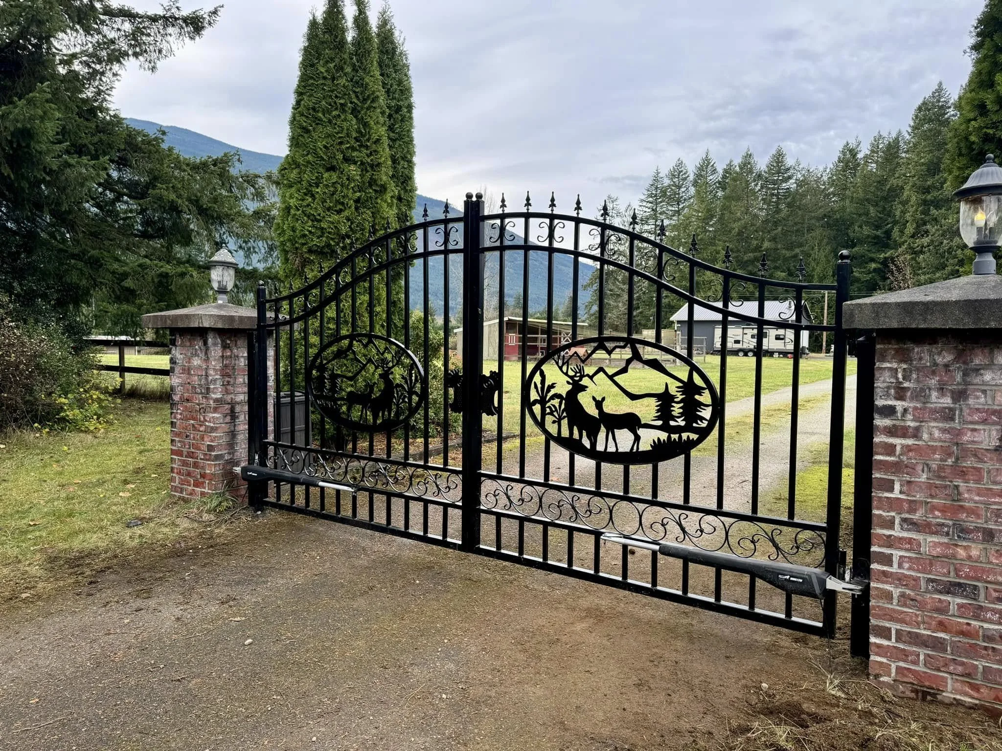 Black wrought iron gate with mountain and deer silhouettes, flanked by brick pillars with lanterns, leading to a grassy yard with trees and mountains in the background.