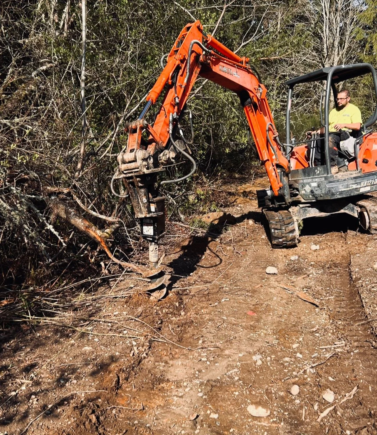 A construction worker operating an orange mini excavator clearing land in a wooded area.