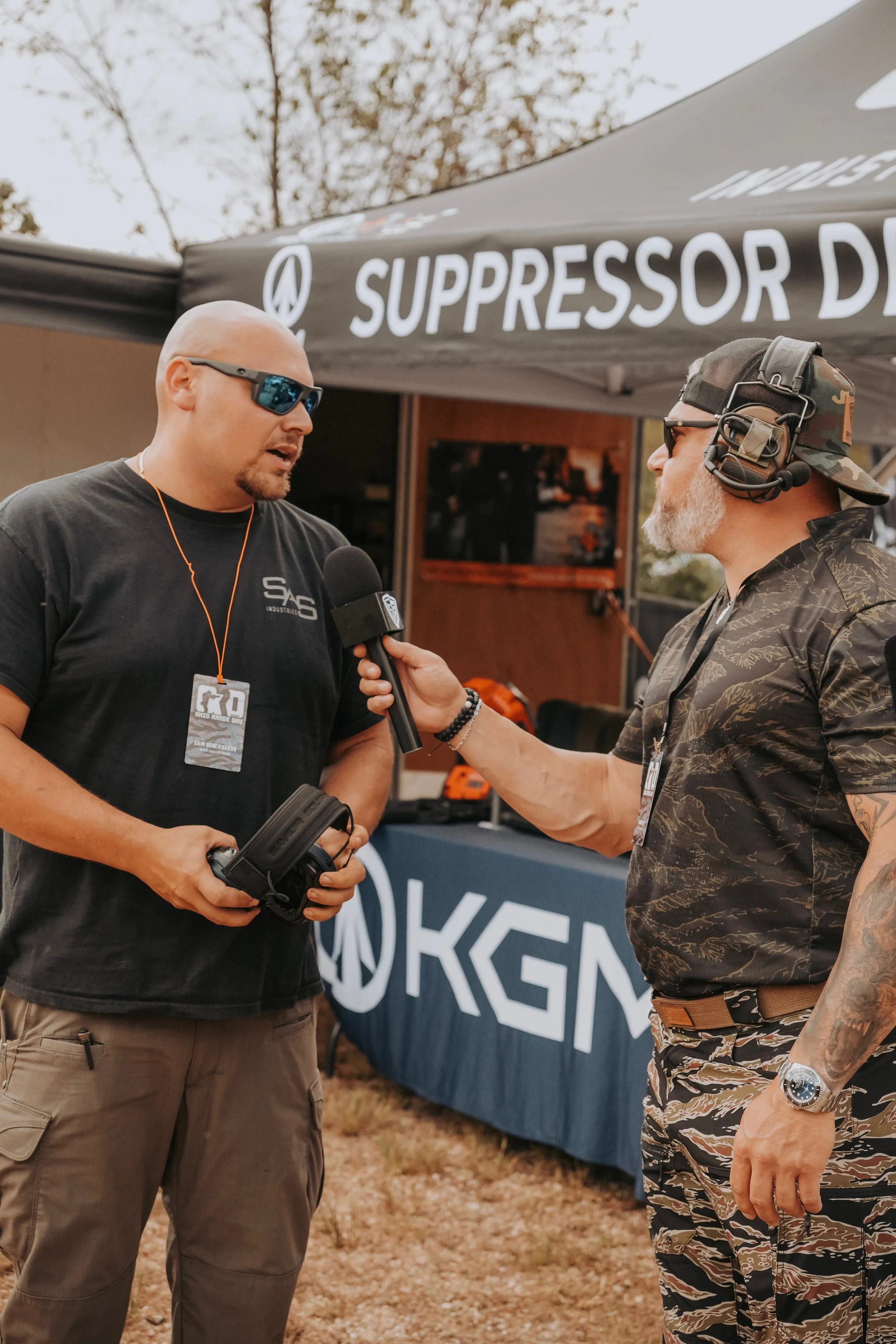 Two men are having an interview outdoors at an event. The man on the left is wearing sunglasses, a black t-shirt, and has a lanyard with a badge. The man on the right is wearing a camouflage cap, sunglasses, headphones, a camouflage t-shirt, and camouflage pants. They are standing near a black tent with the words 'SUPPRESSOR D' visible and a table with display items.