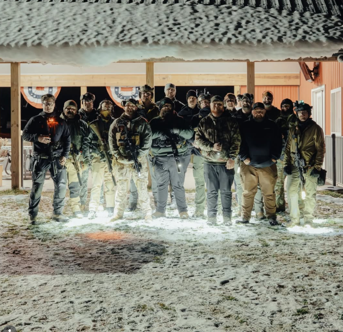 Group of armed men standing together outdoors at night, some holding guns, under a canopy decorated with patriotic bunting.
